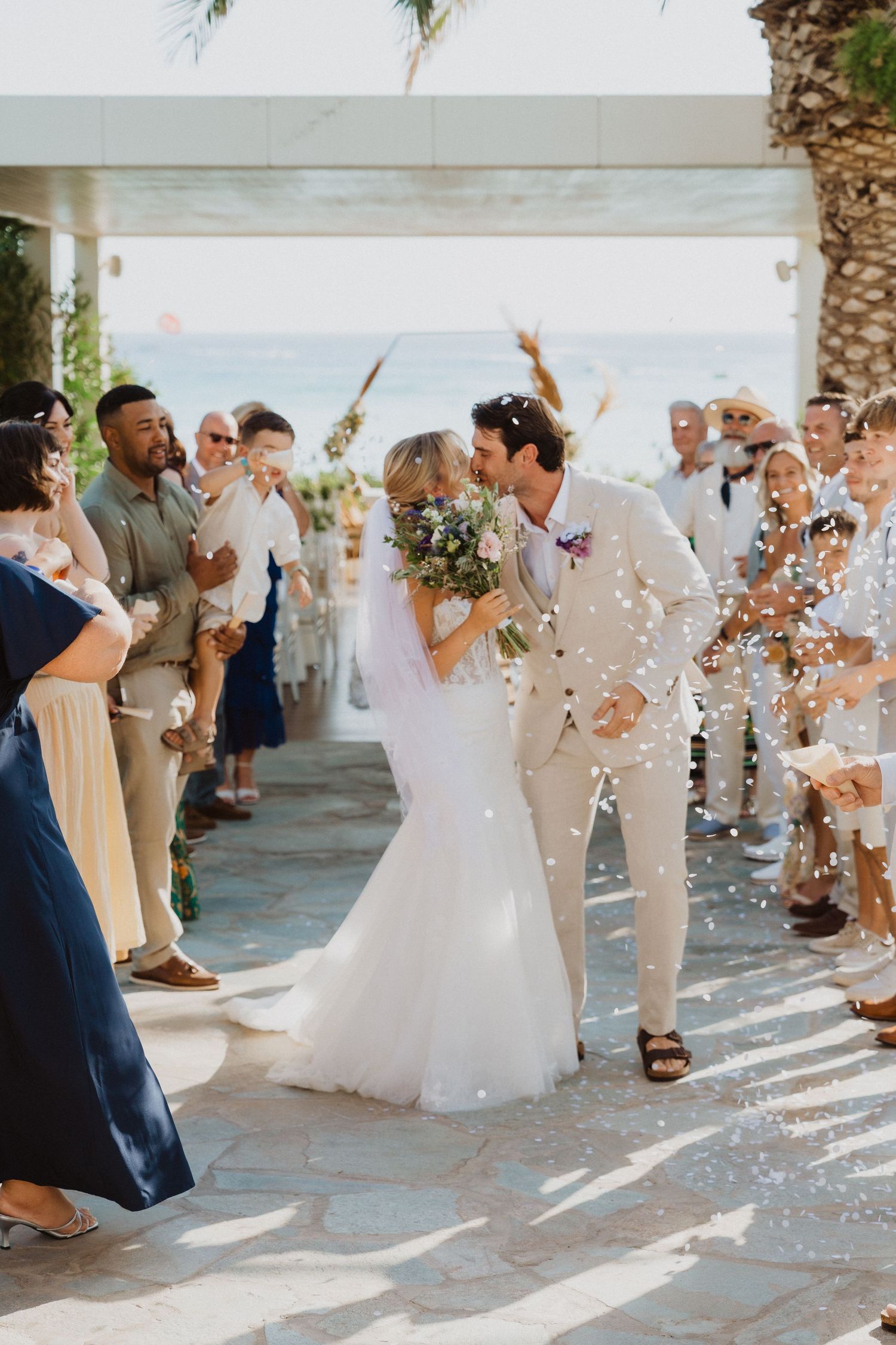 Wedding guests toss confetti at a tropical outdoor celebration with palm trees and bright sunlight.