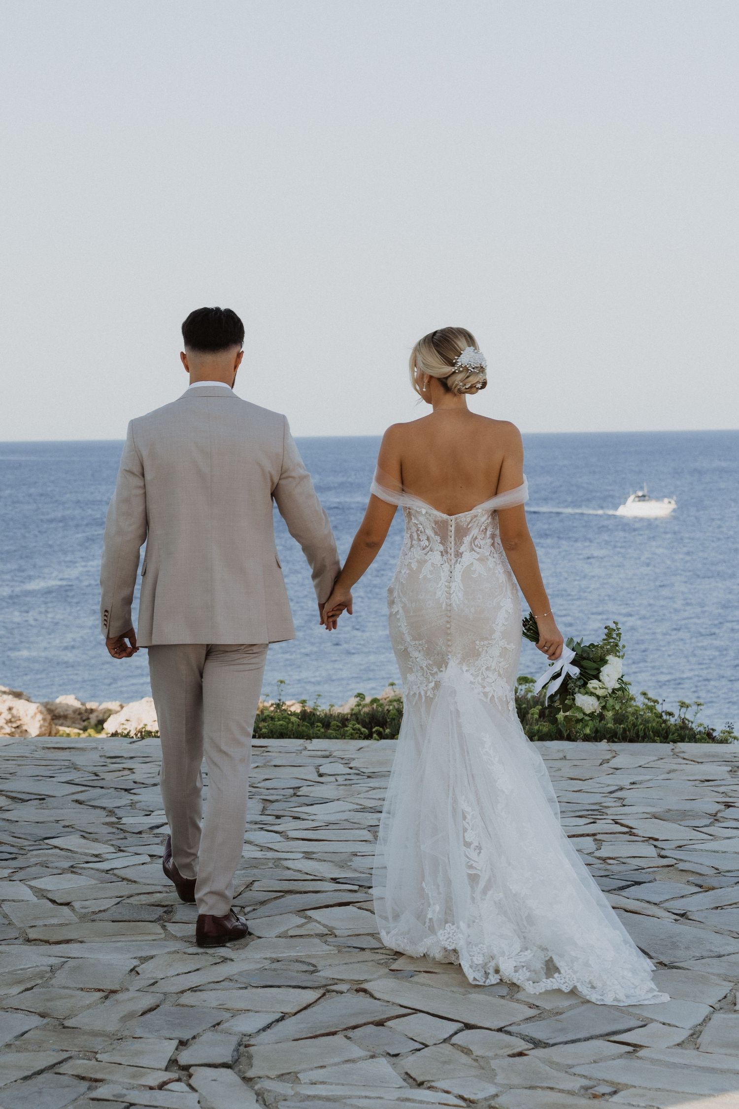 Newlyweds walk along a stone pathway overlooking the Mediterranean Sea.