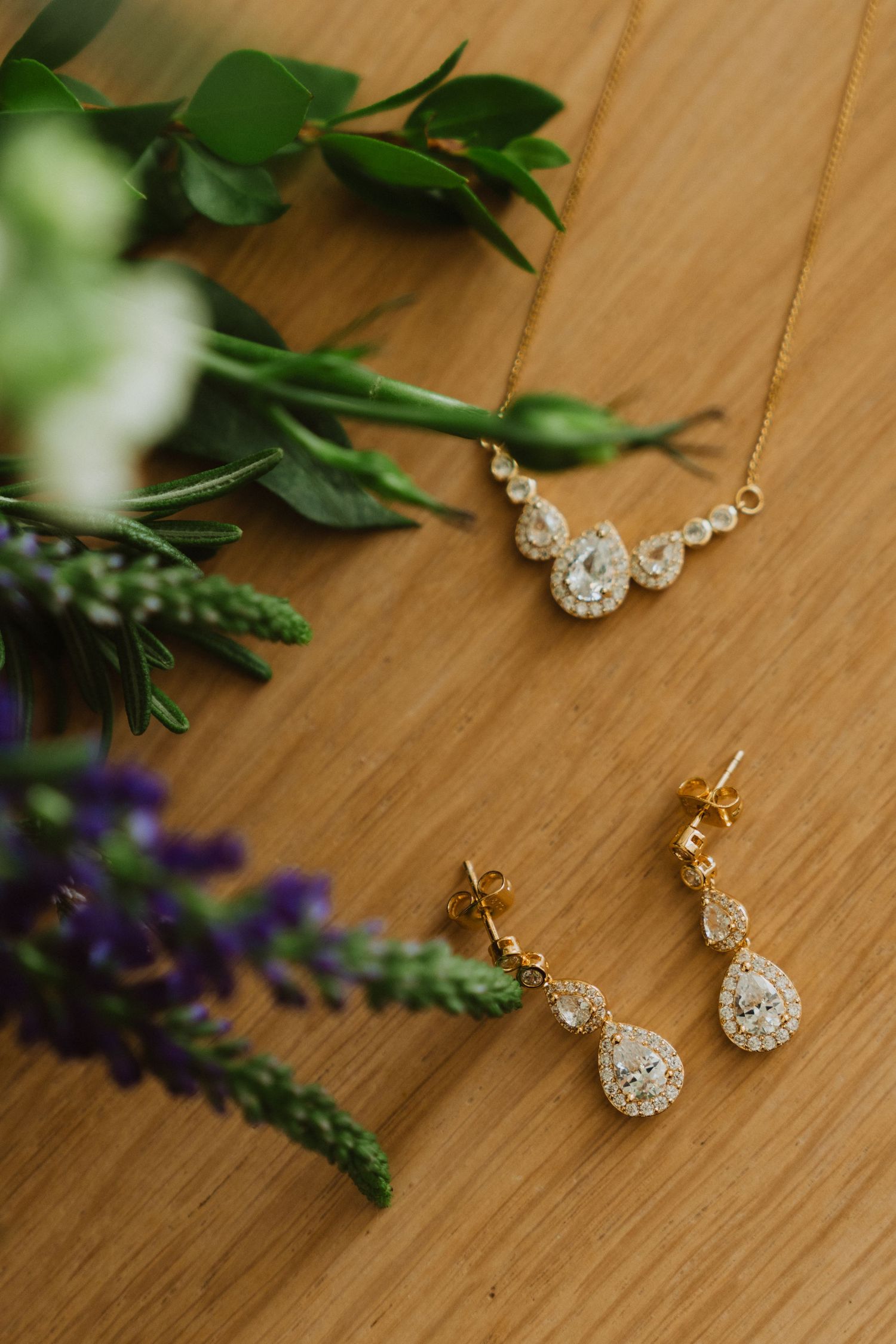 Elegant diamond drop earrings and necklace displayed with purple flowers and greenery on wooden surface.