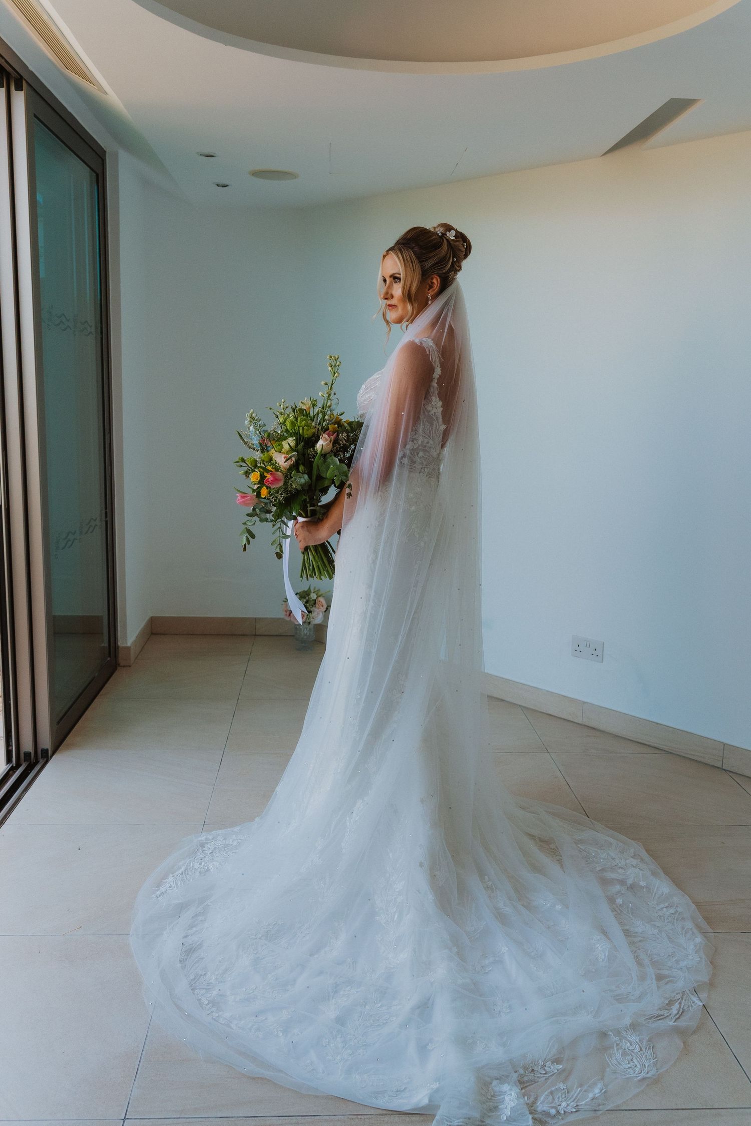 A bride in a flowing white lace wedding gown with a long veil stands in an elegant hallway.