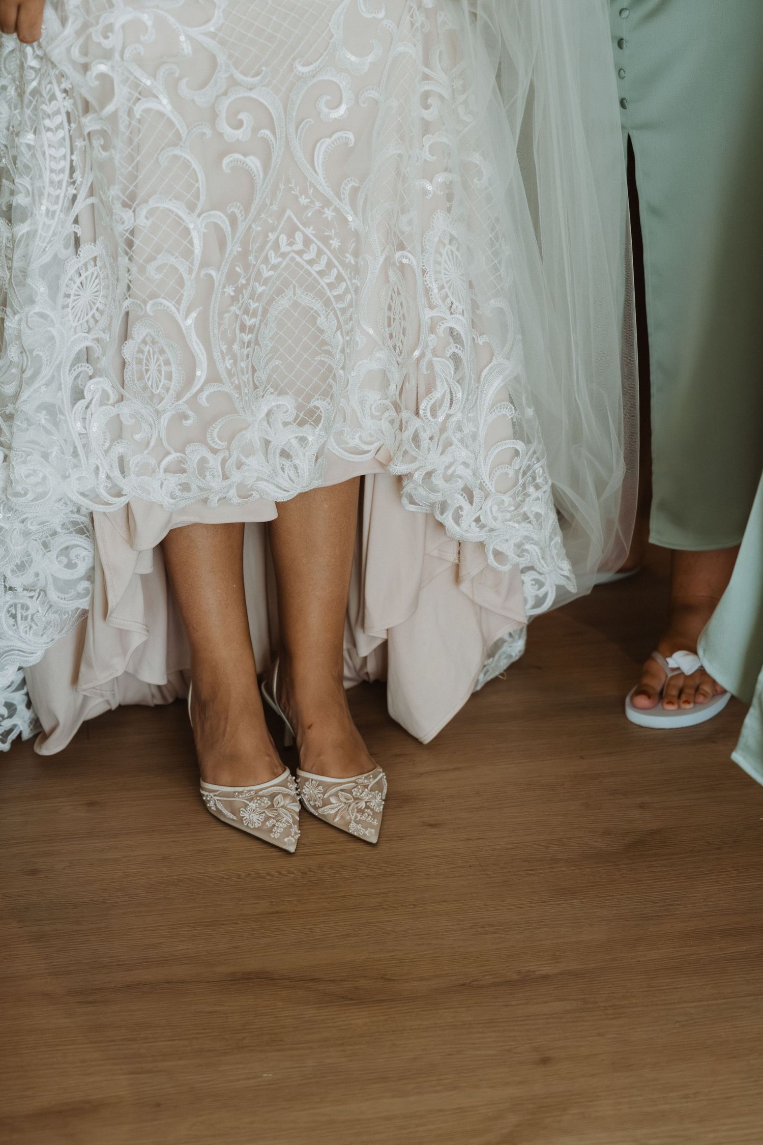 Close-up view of elegant lace wedding dress hem and bridal shoes on wooden floor.