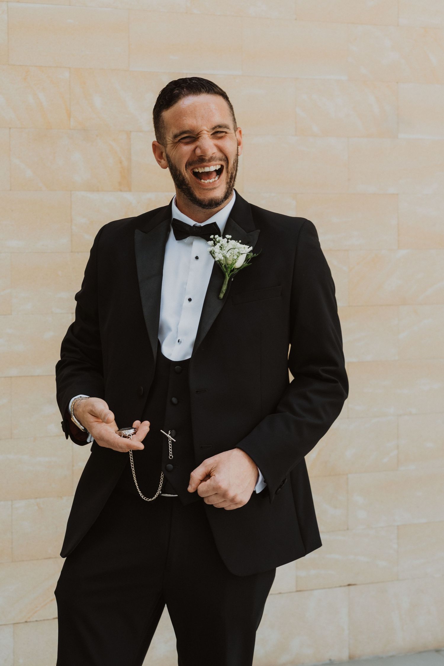 A groom in a black tuxedo and bowtie laughs while getting ready on his wedding day.