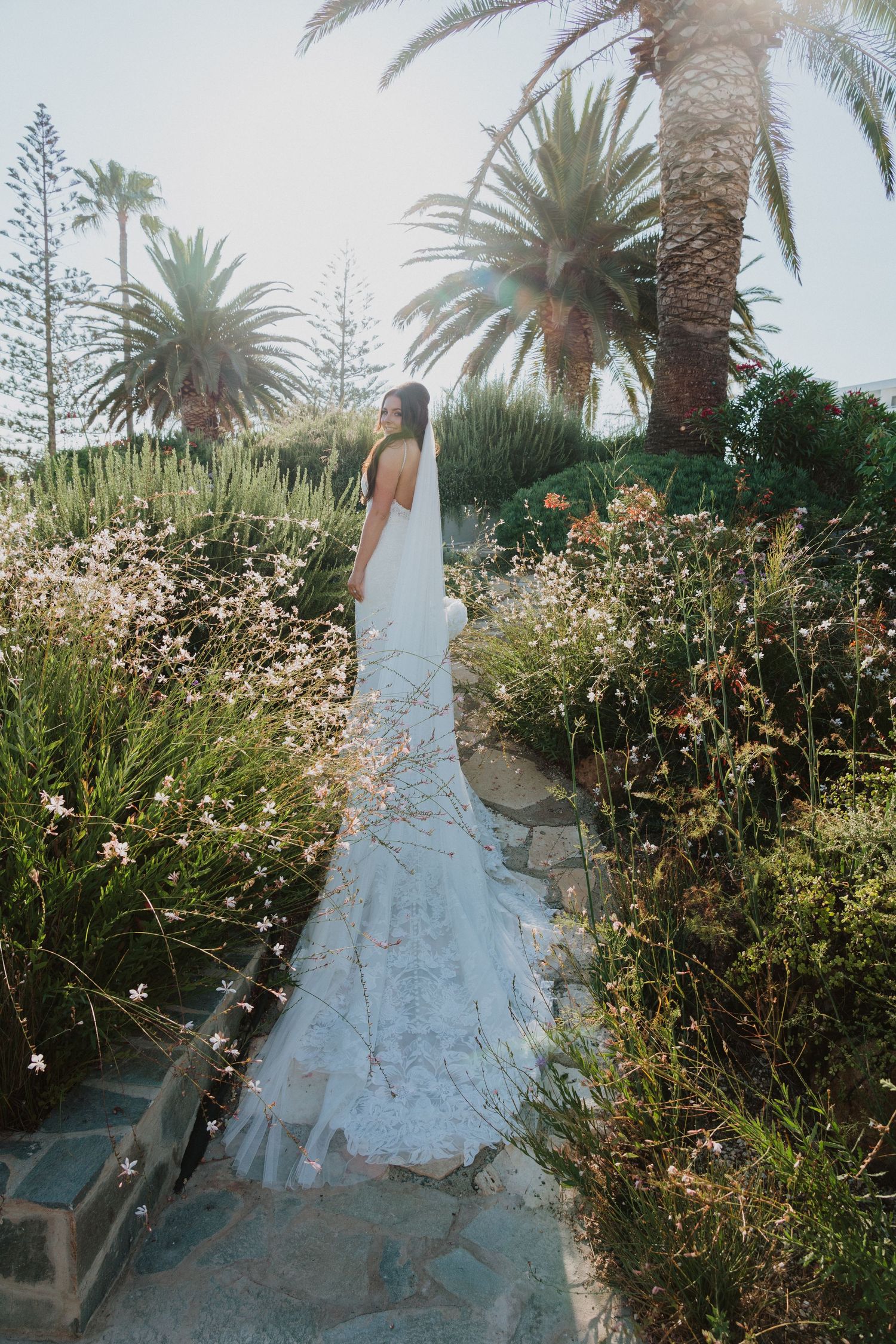 A bride in a flowing white lace dress walks along a garden path surrounded by palm trees and wildflowers.