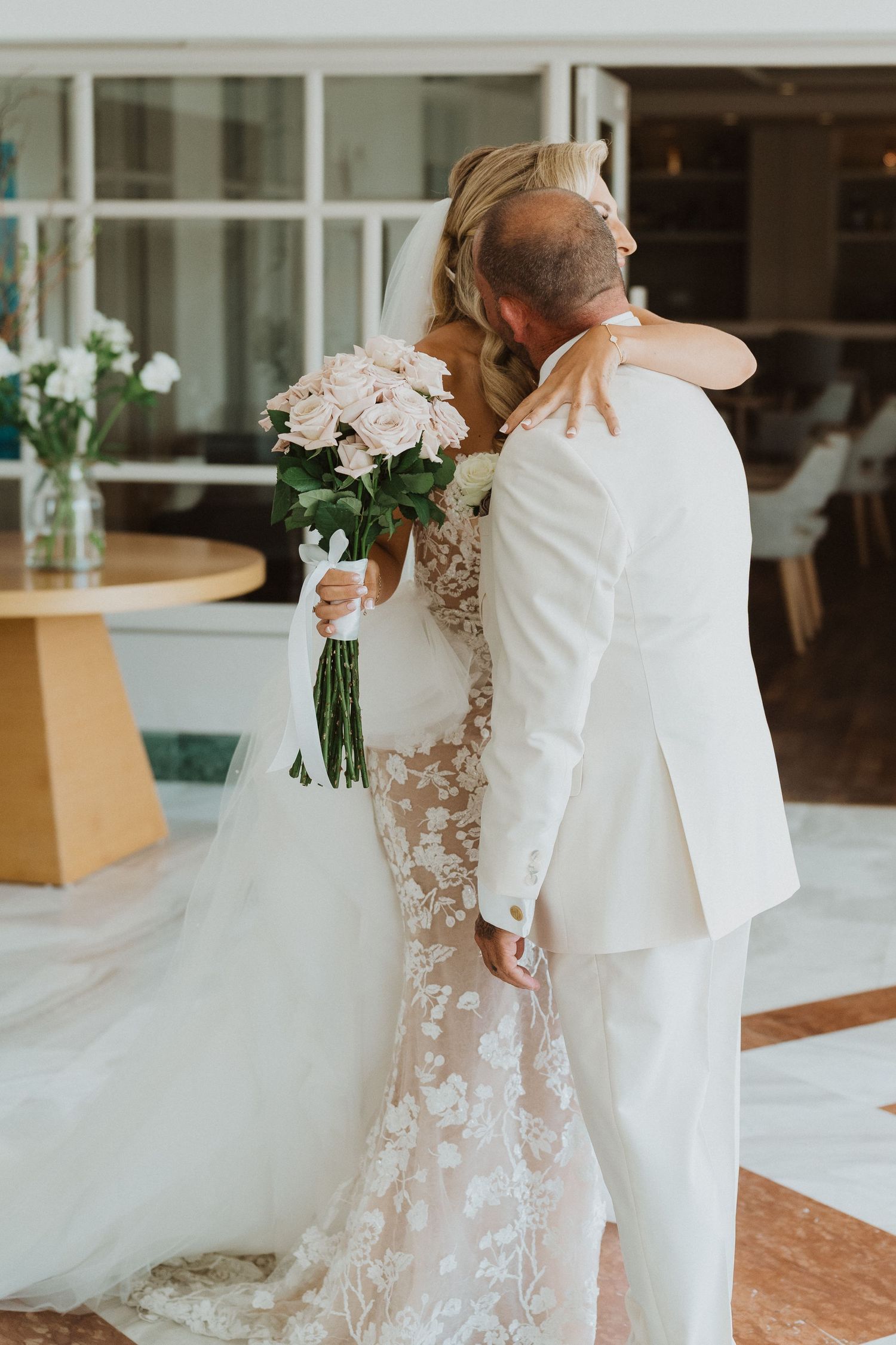 A tender wedding moment as a couple embraces while the bride holds a bouquet of pink roses in her lace gown.