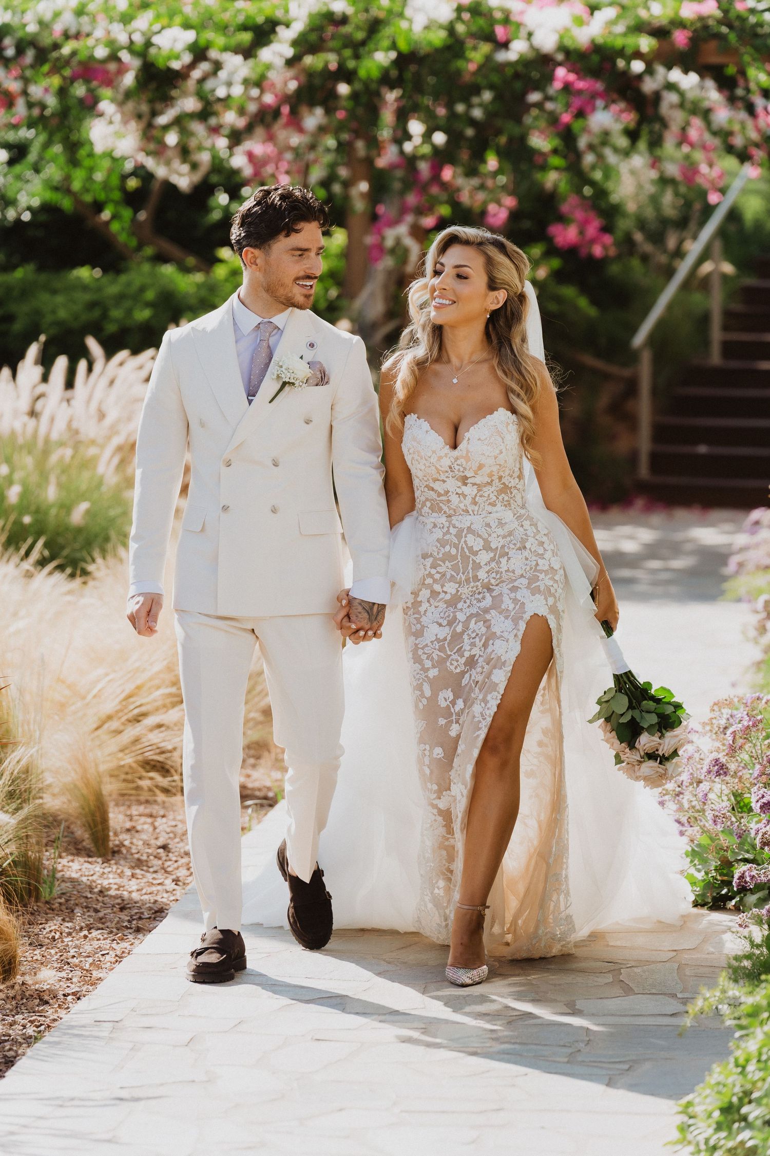 A couple walking hand in hand on their wedding day, wearing a white double-breasted suit and floral lace gown in a garden setting.