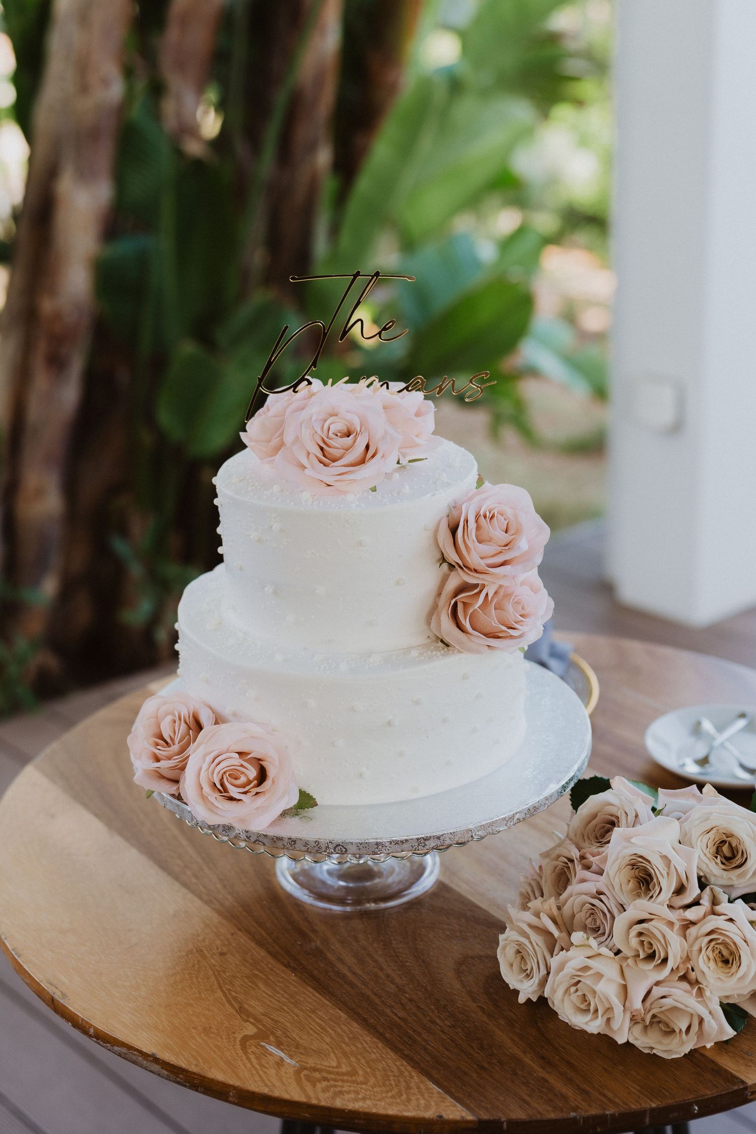 Simple white wedding cake decorated with blush pink roses sits on a wooden cake stand in a rustic setting.