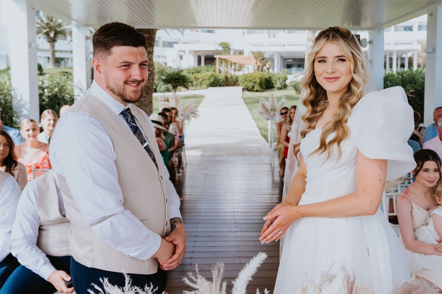 A couple in wedding attire stand together at an outdoor ceremony with guests seated behind them.