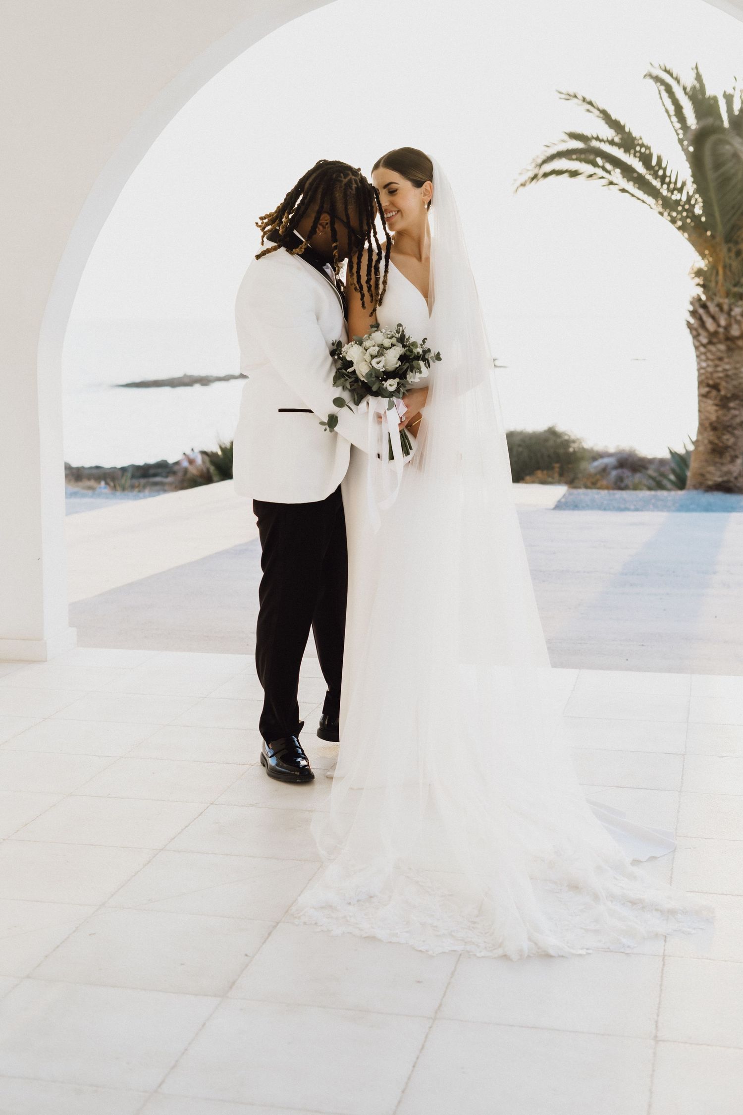Wedding couple embraces under white Mediterranean archway with ocean view.