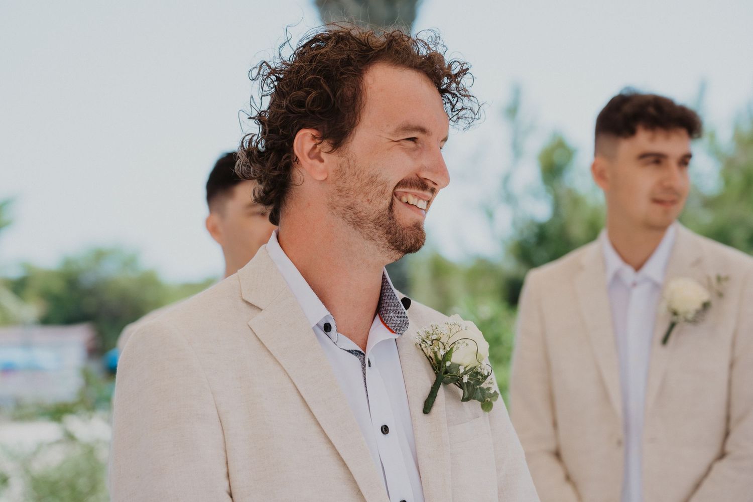 Wedding party member in beige suit with boutonniere smiles during outdoor ceremony.
