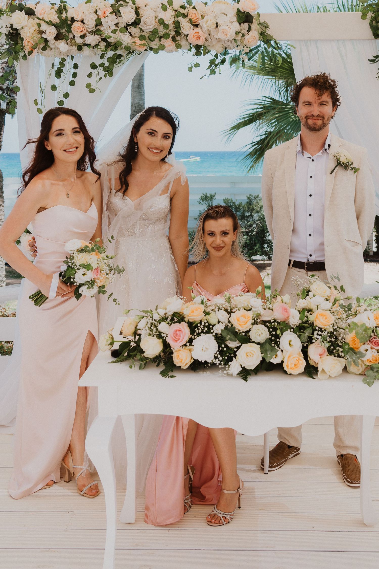 Wedding party poses in front of white floral arch with ocean view and palm trees in background.