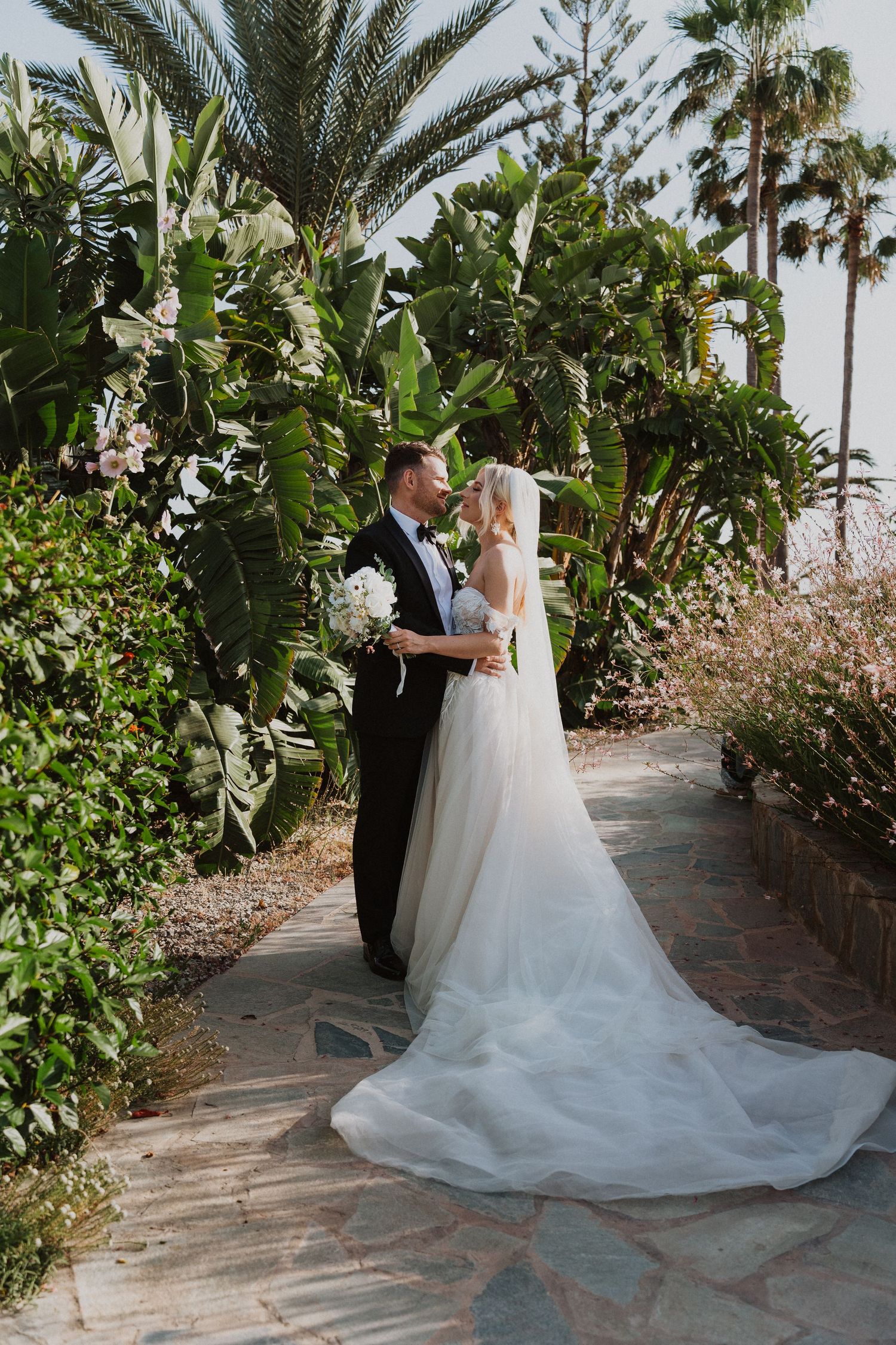 A romantic wedding portrait amid lush tropical gardens with palm trees and stone pathway.
