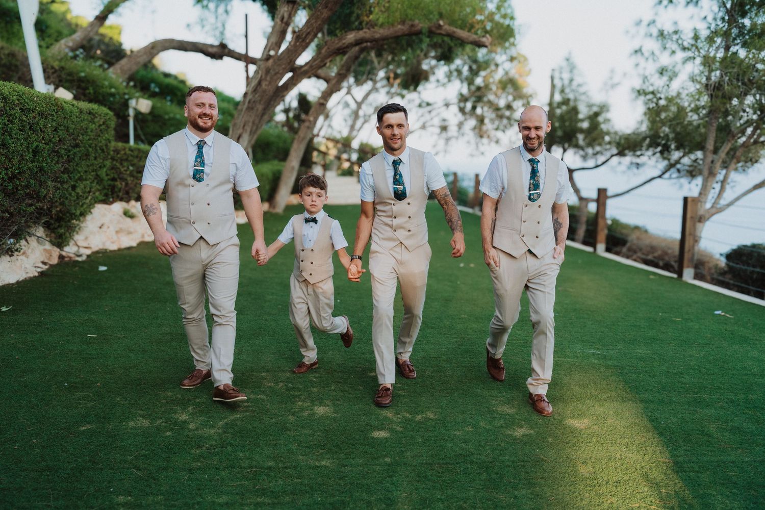 Wedding party members in beige suits walk together on grass with ocean view.