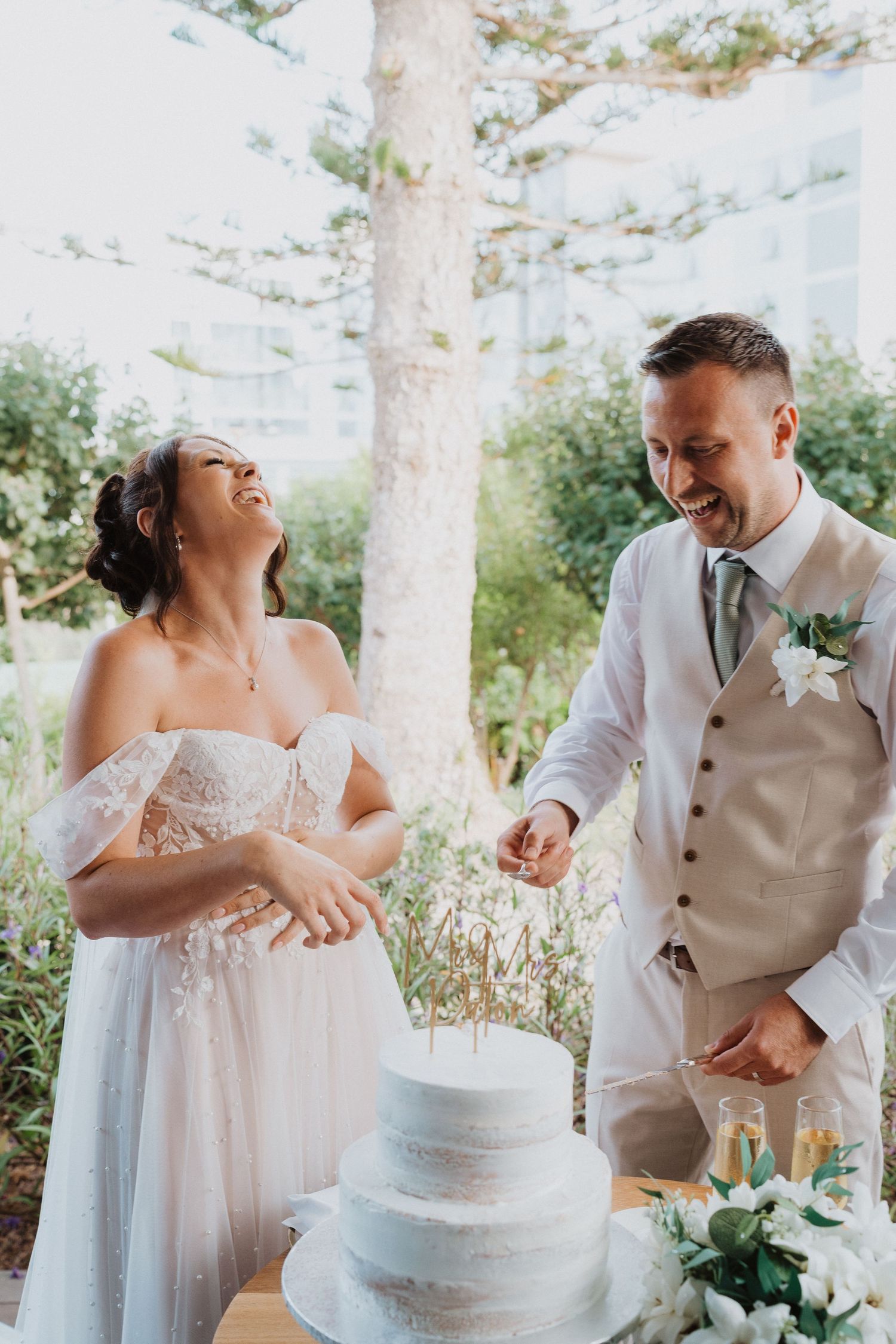 Newlyweds share a joyful moment while cutting their elegant white wedding cake outdoors under tall pine trees.