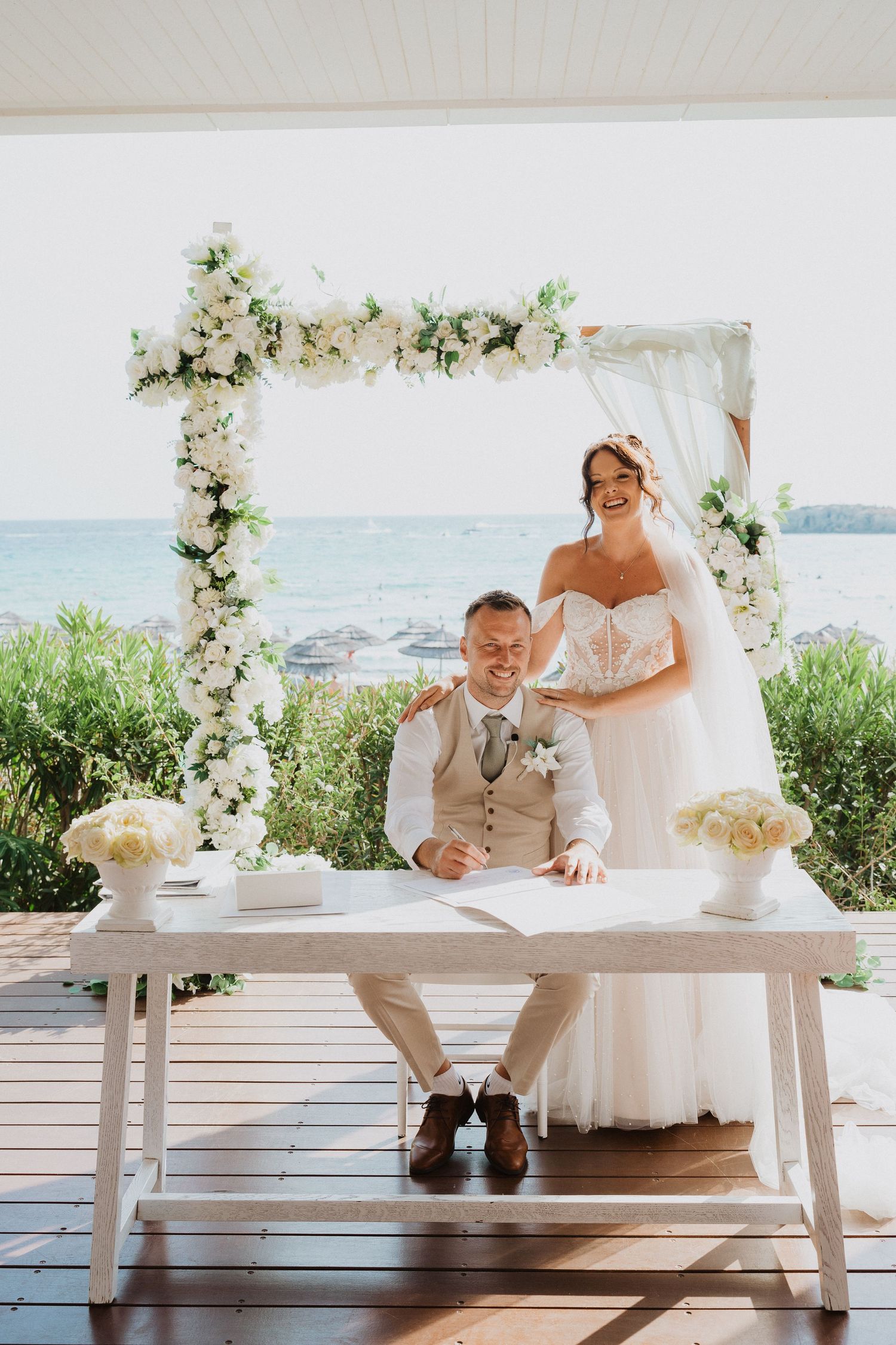 A wedding couple signs documents at an elegant white floral arch overlooking the ocean during their outdoor ceremony.