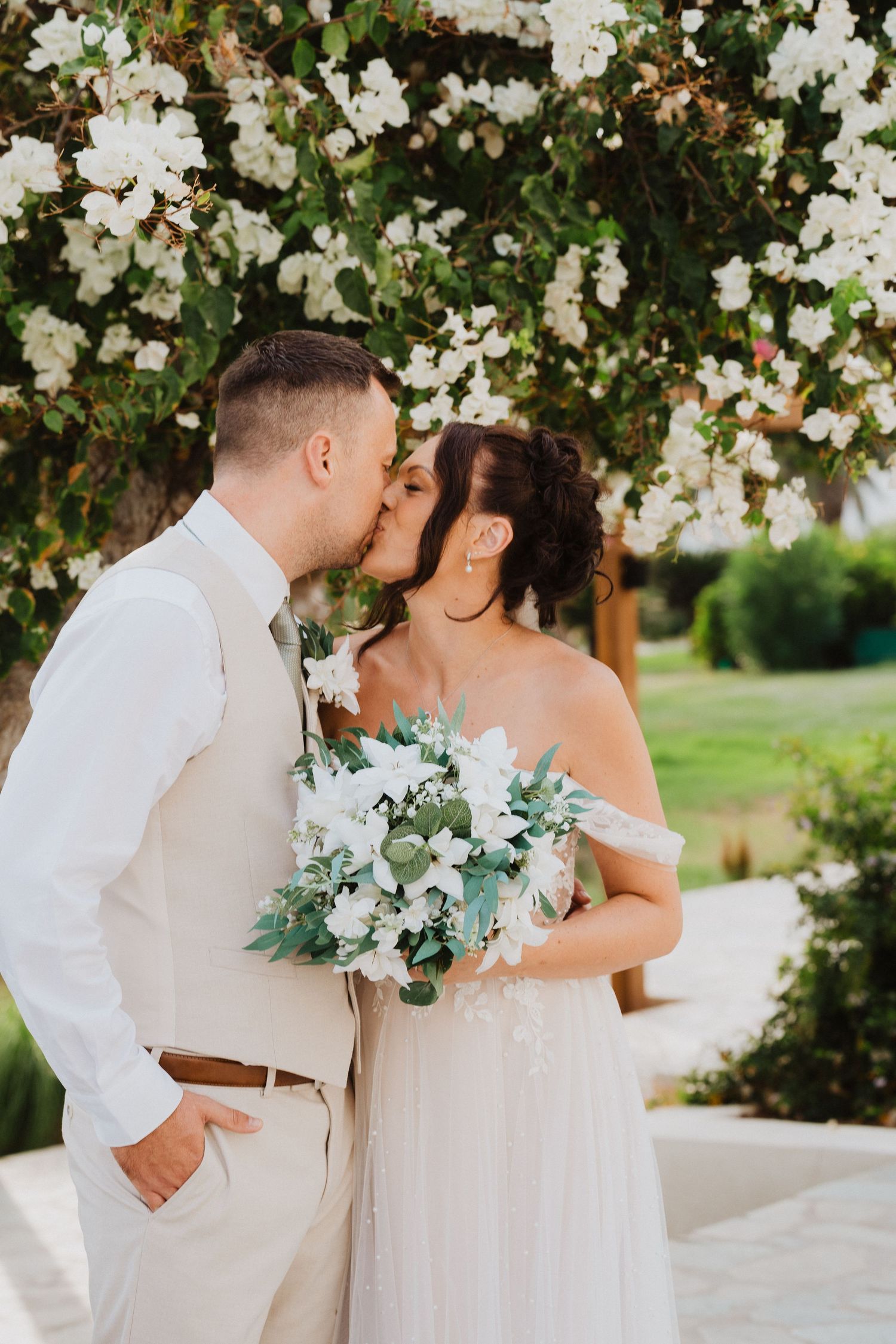 A romantic wedding couple shares a kiss in front of blooming white flowers on a summer day.