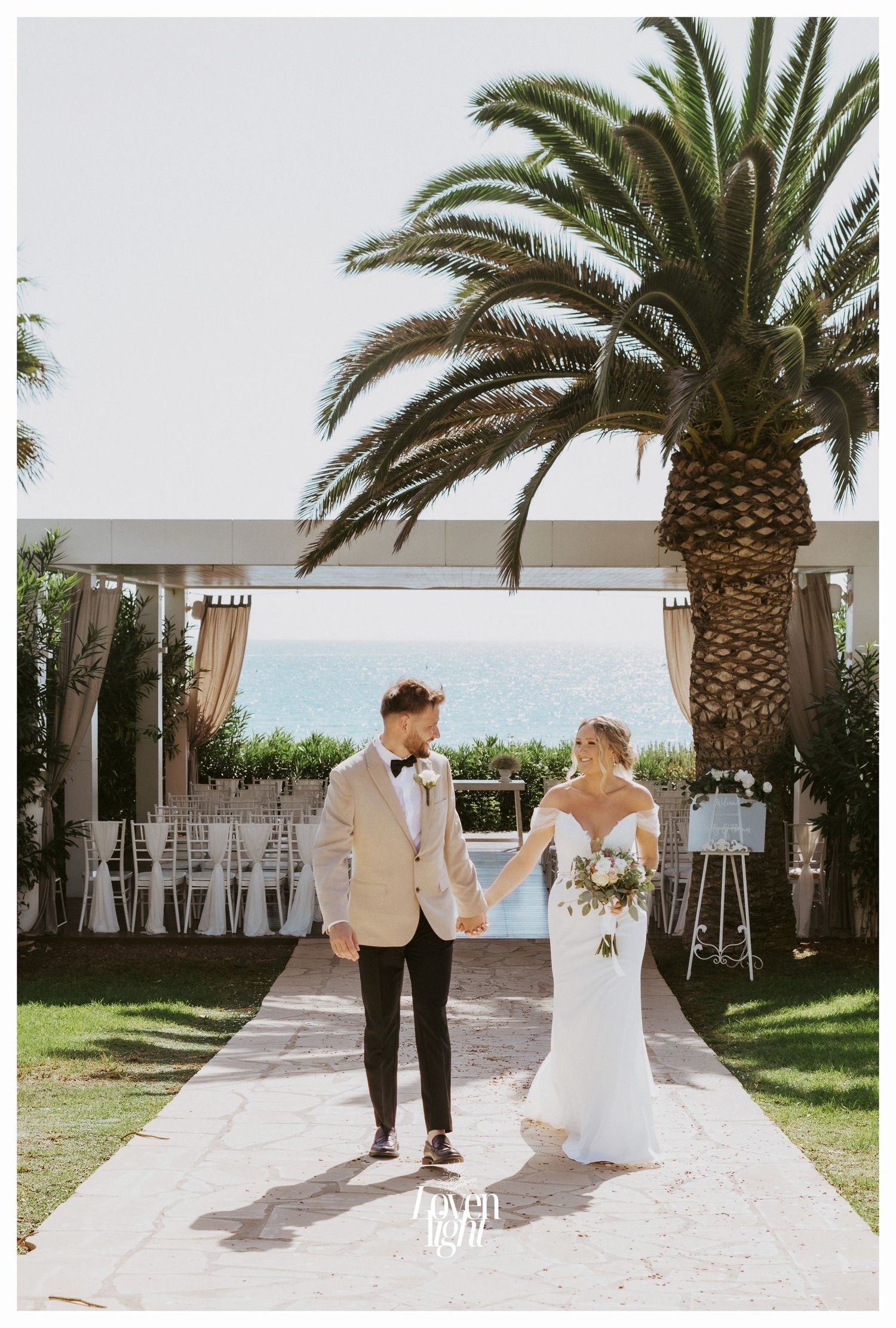 A wedding couple poses on a white pathway with tropical palm trees and ocean views.