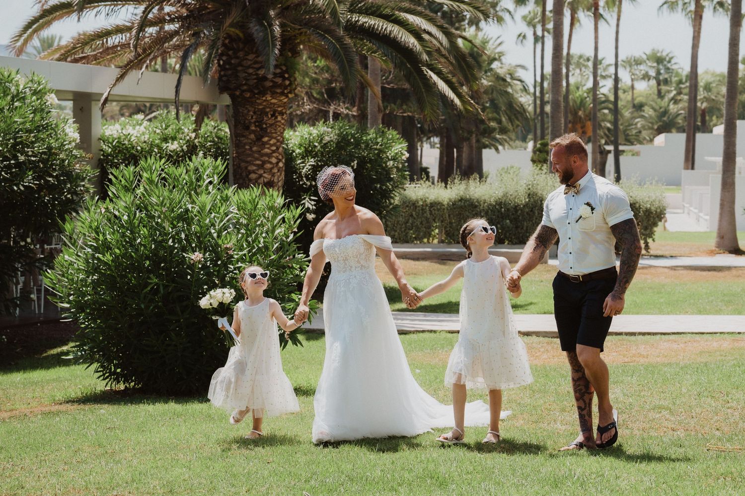 Family in white attire holds hands while walking along a palm-lined garden path.