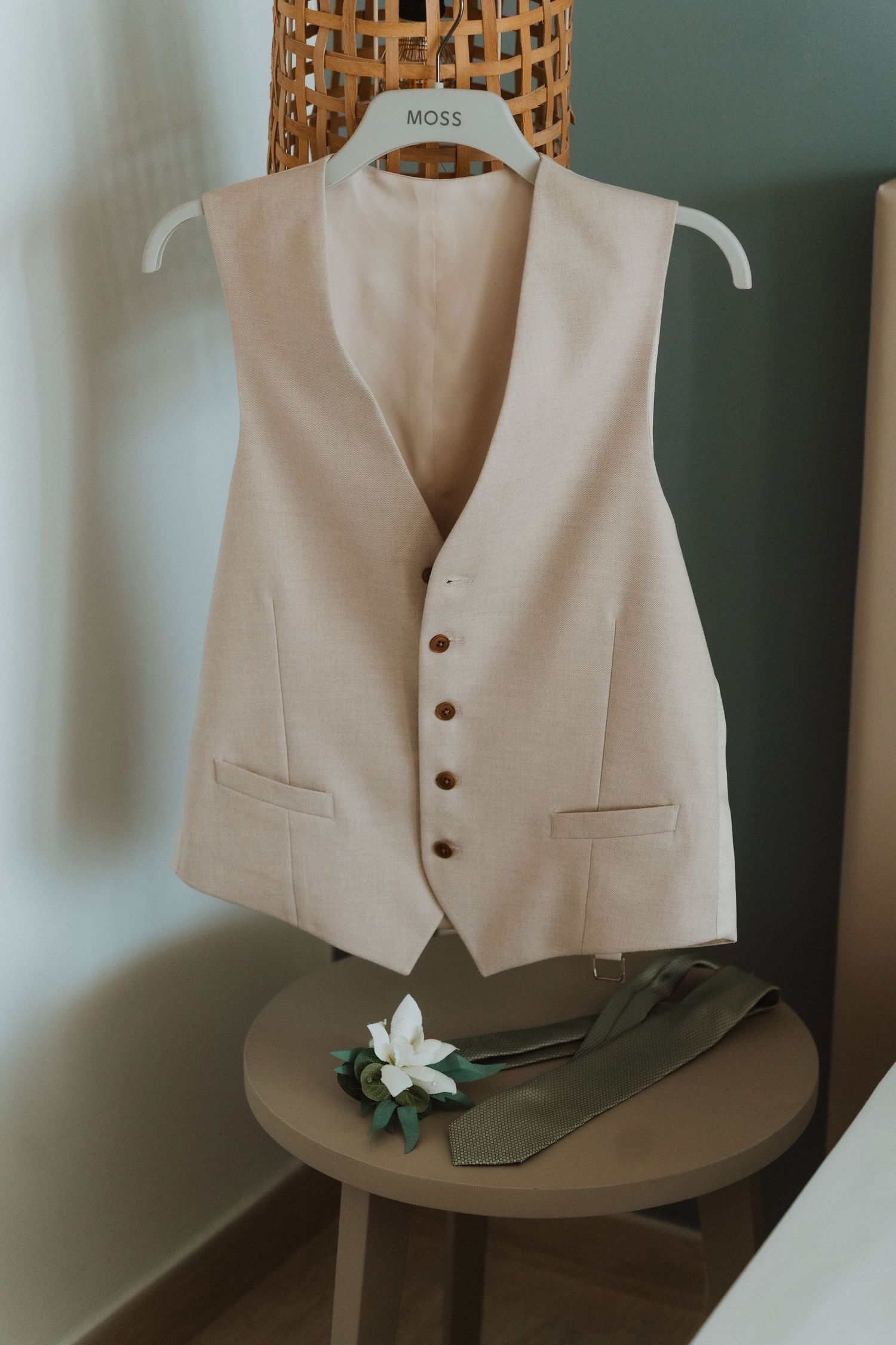 Elegant cream waistcoat with brass buttons hangs on wooden hanger above table with white flower detail.