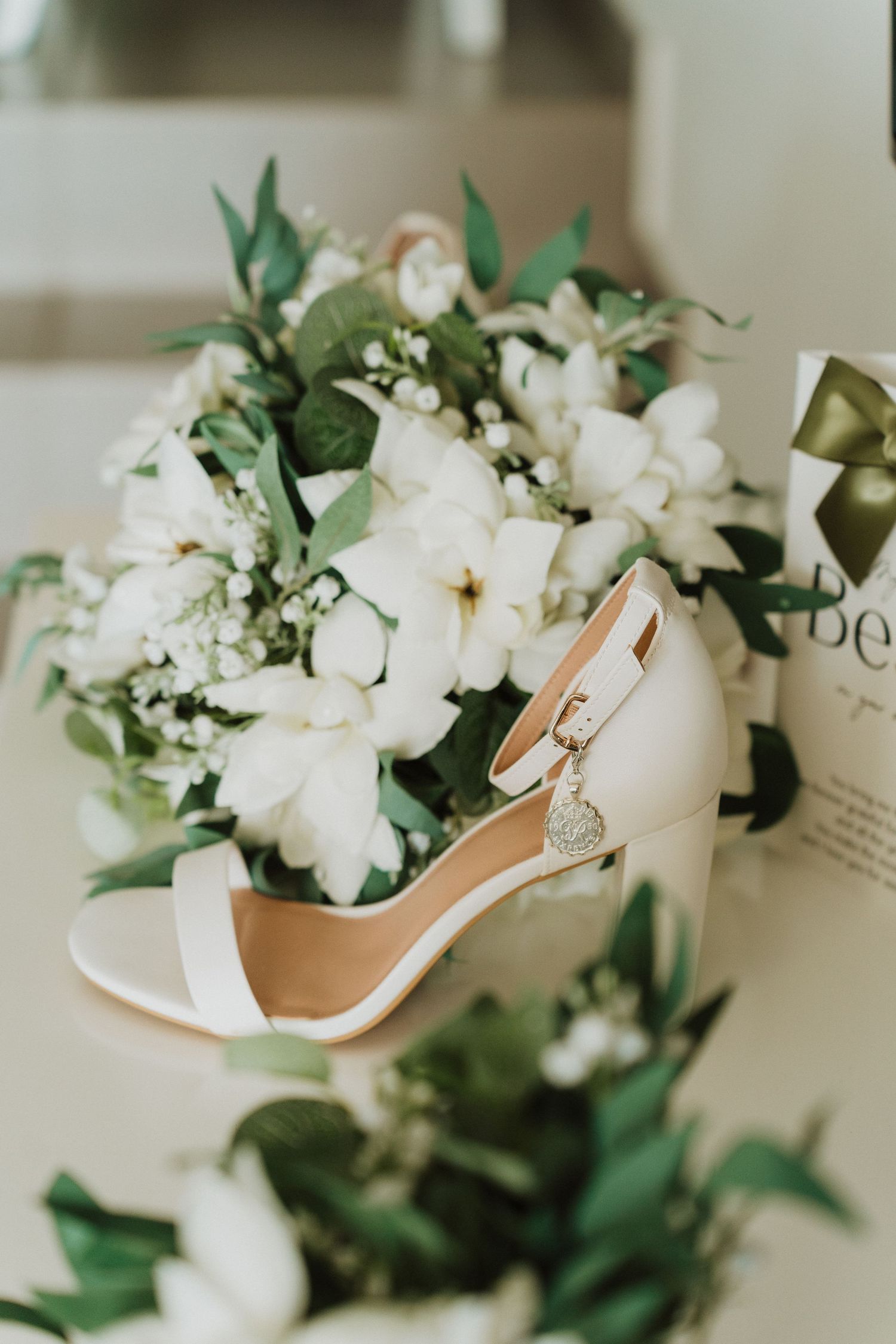 Elegant white bridal heel sandal surrounded by delicate white flowers and green foliage arrangements on cream surface.