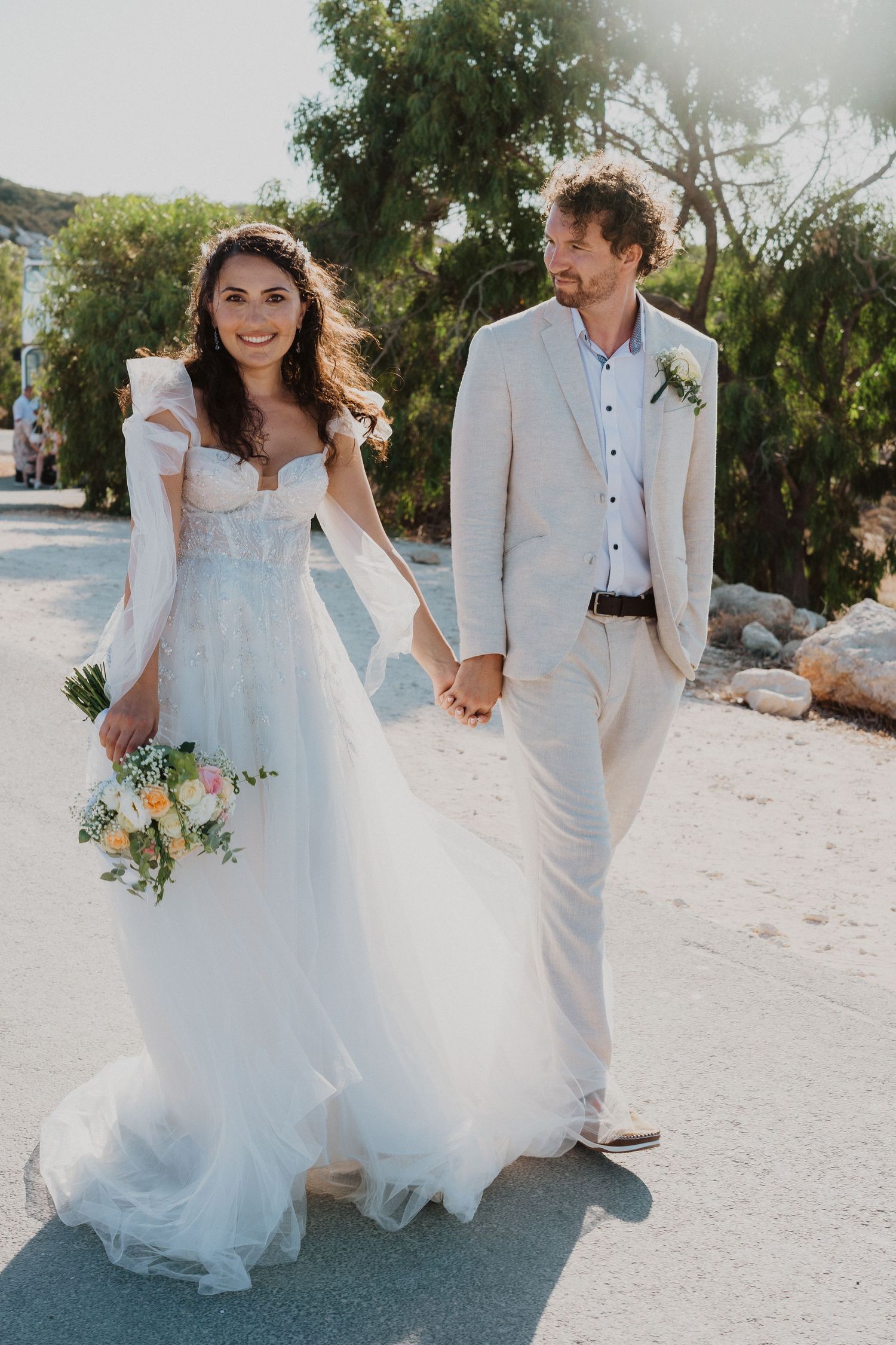 Wedding couple in white attire walking hand in hand outdoors on a sunny day.