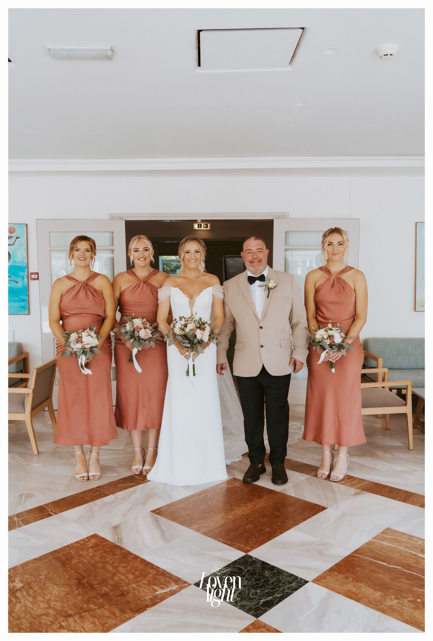 Wedding party poses on black and white checkered floor with bridesmaids in coral dresses and groomsmen in beige suit.