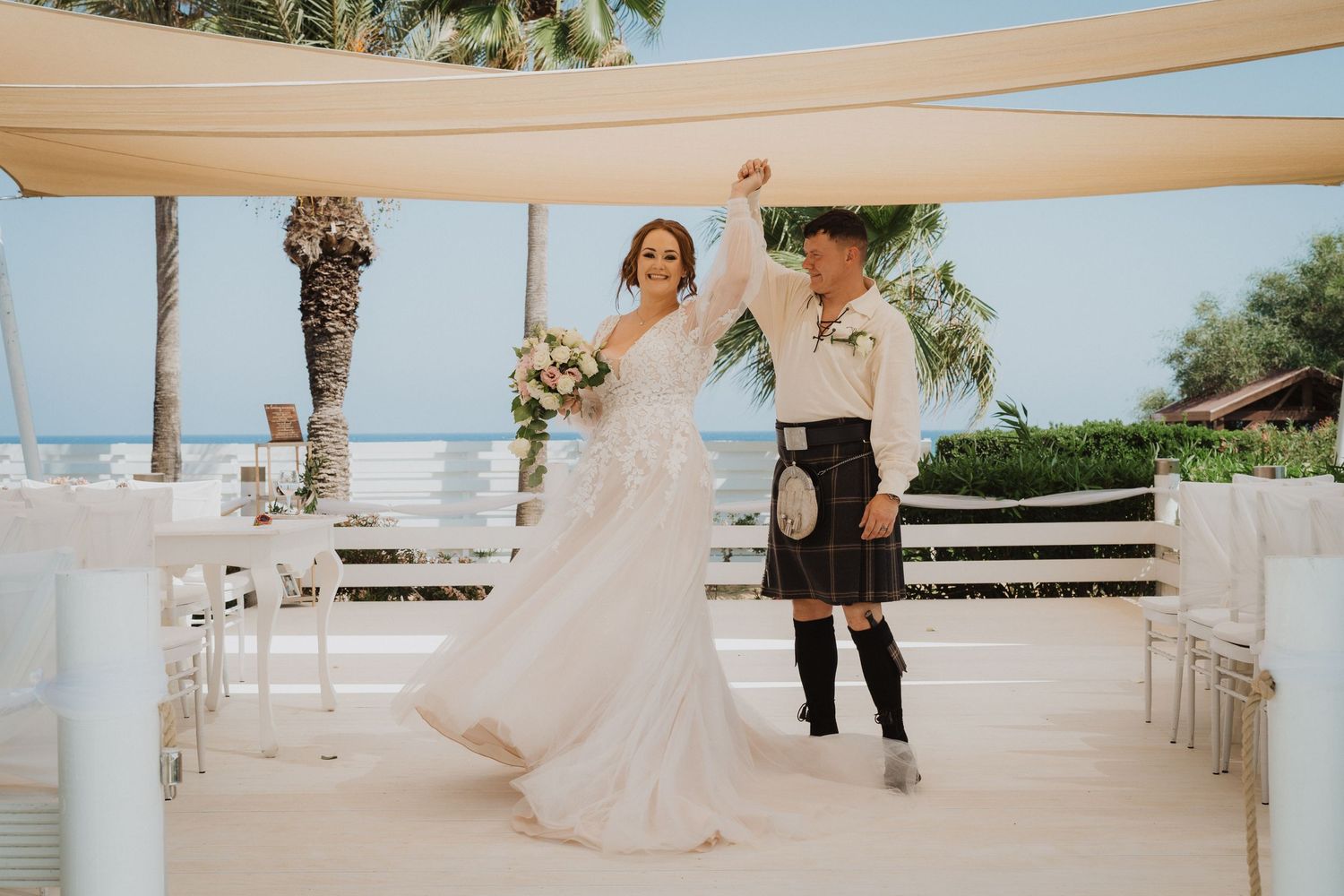 Wedding couple dancing on waterfront terrace under beige canopies with palm trees in background.