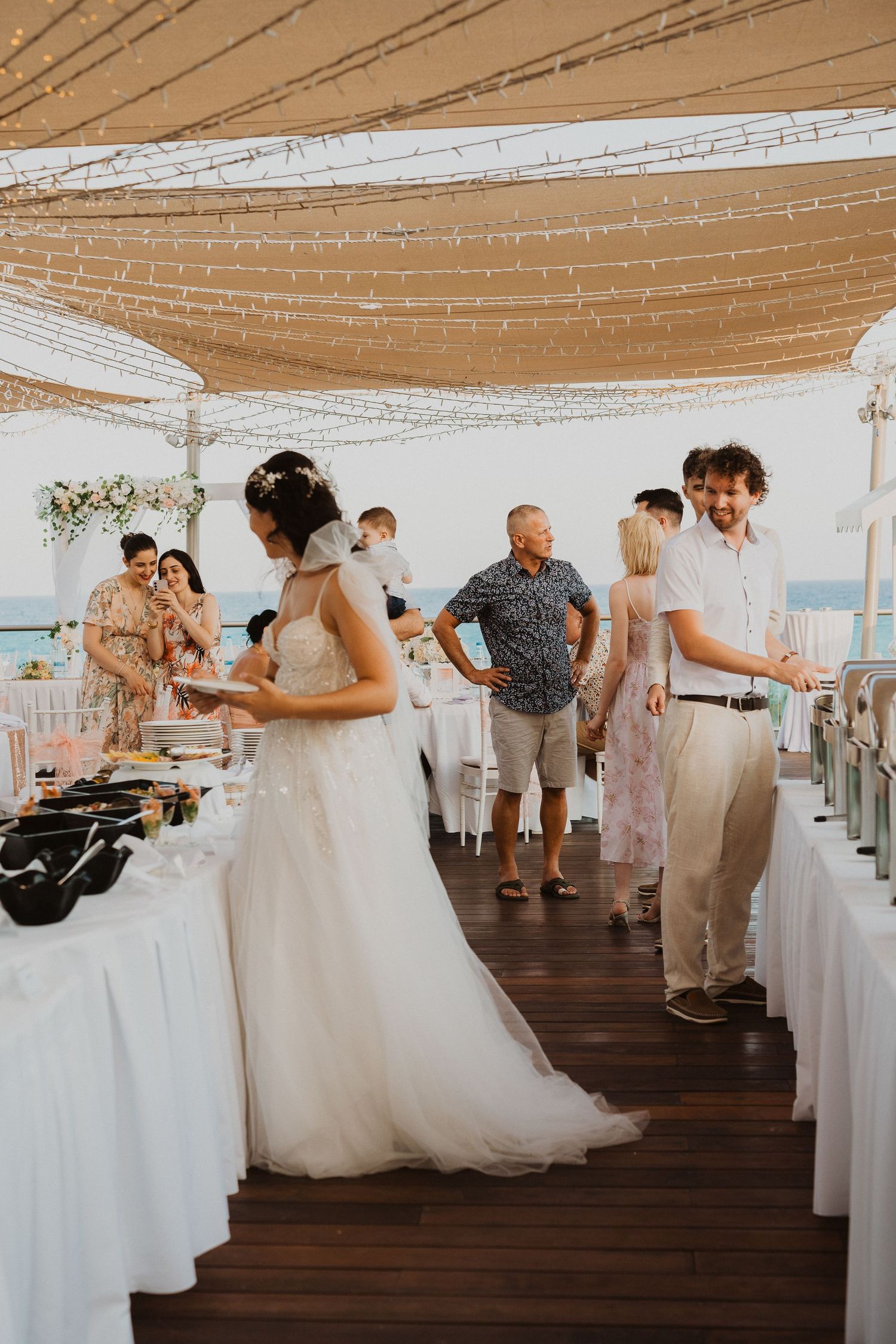 Wedding reception buffet setup with white linens and string lights overhead at beachfront venue.