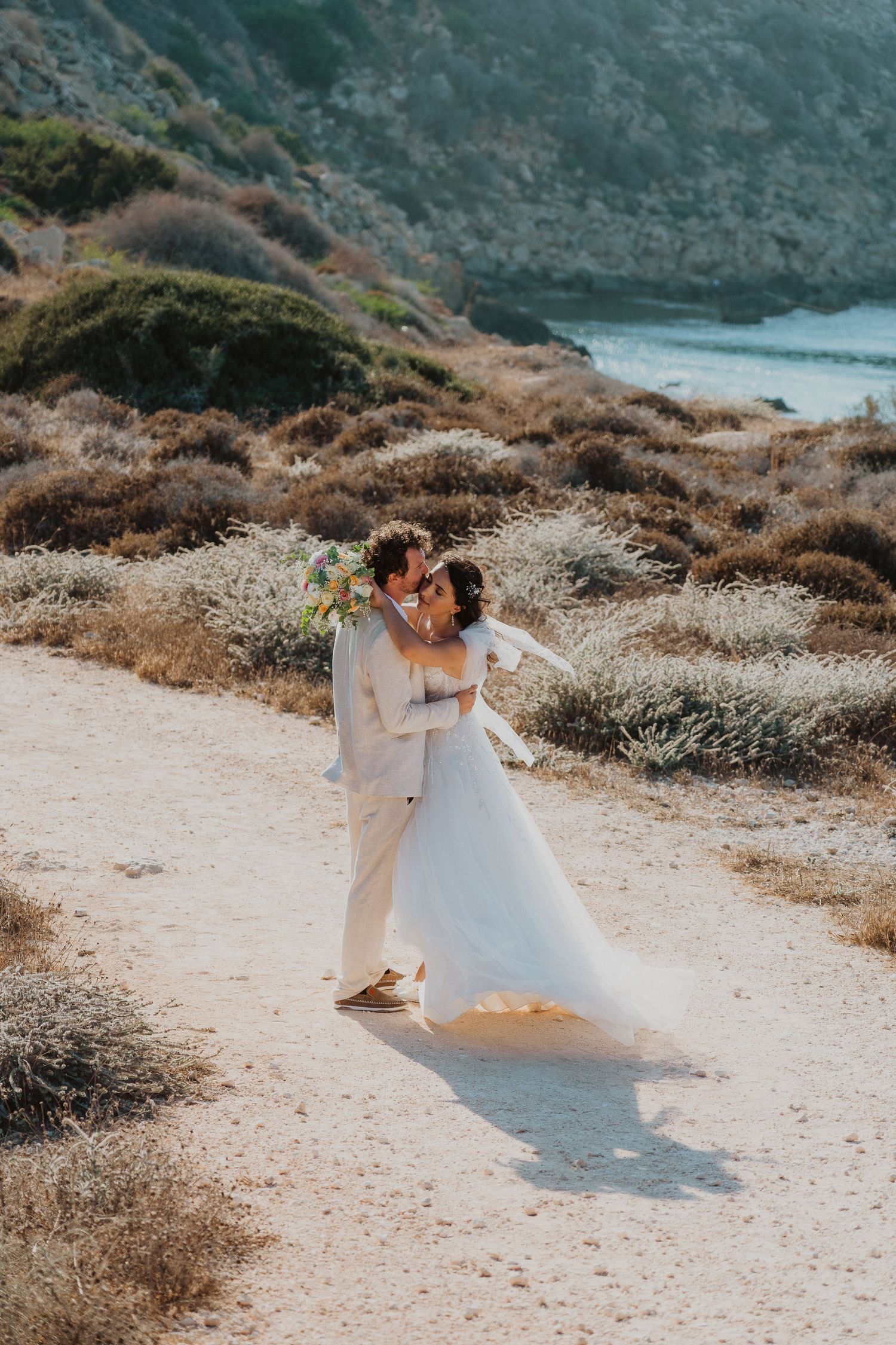 A wedding couple embraces on a scenic coastal trail overlooking blue waters and rugged cliffs.