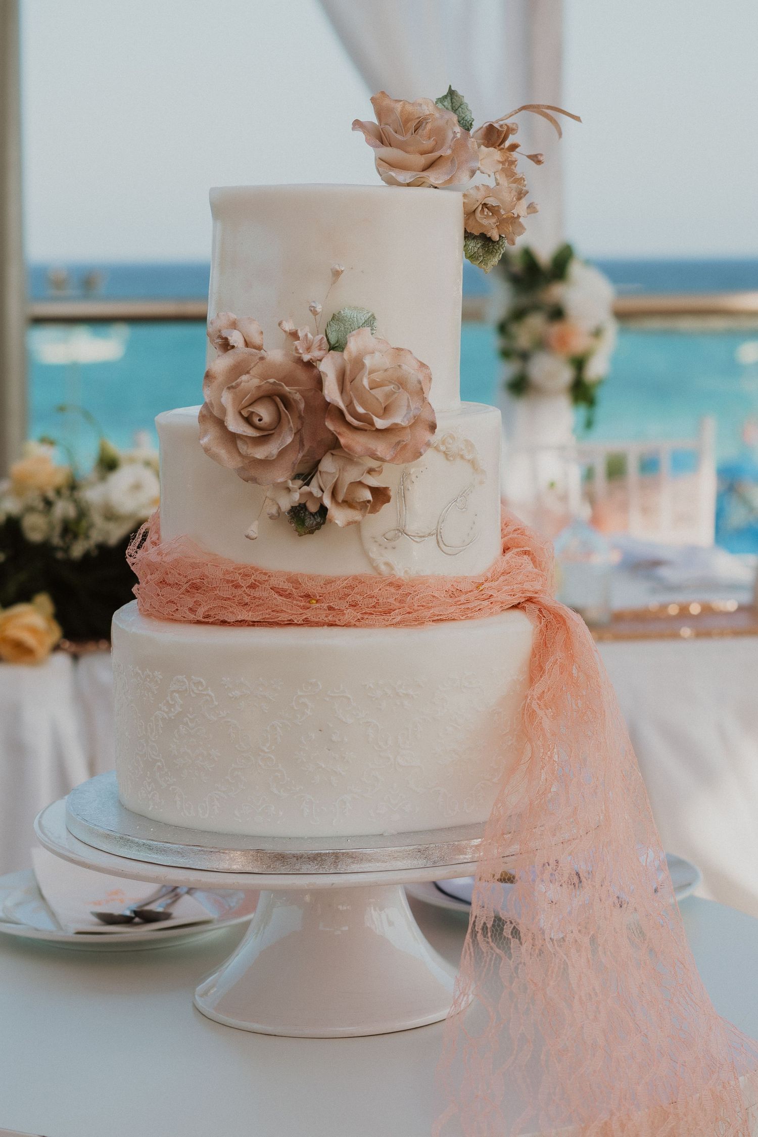 Elegant three-tier white wedding cake adorned with peach roses and flowing tulle ribbon against an ocean backdrop.