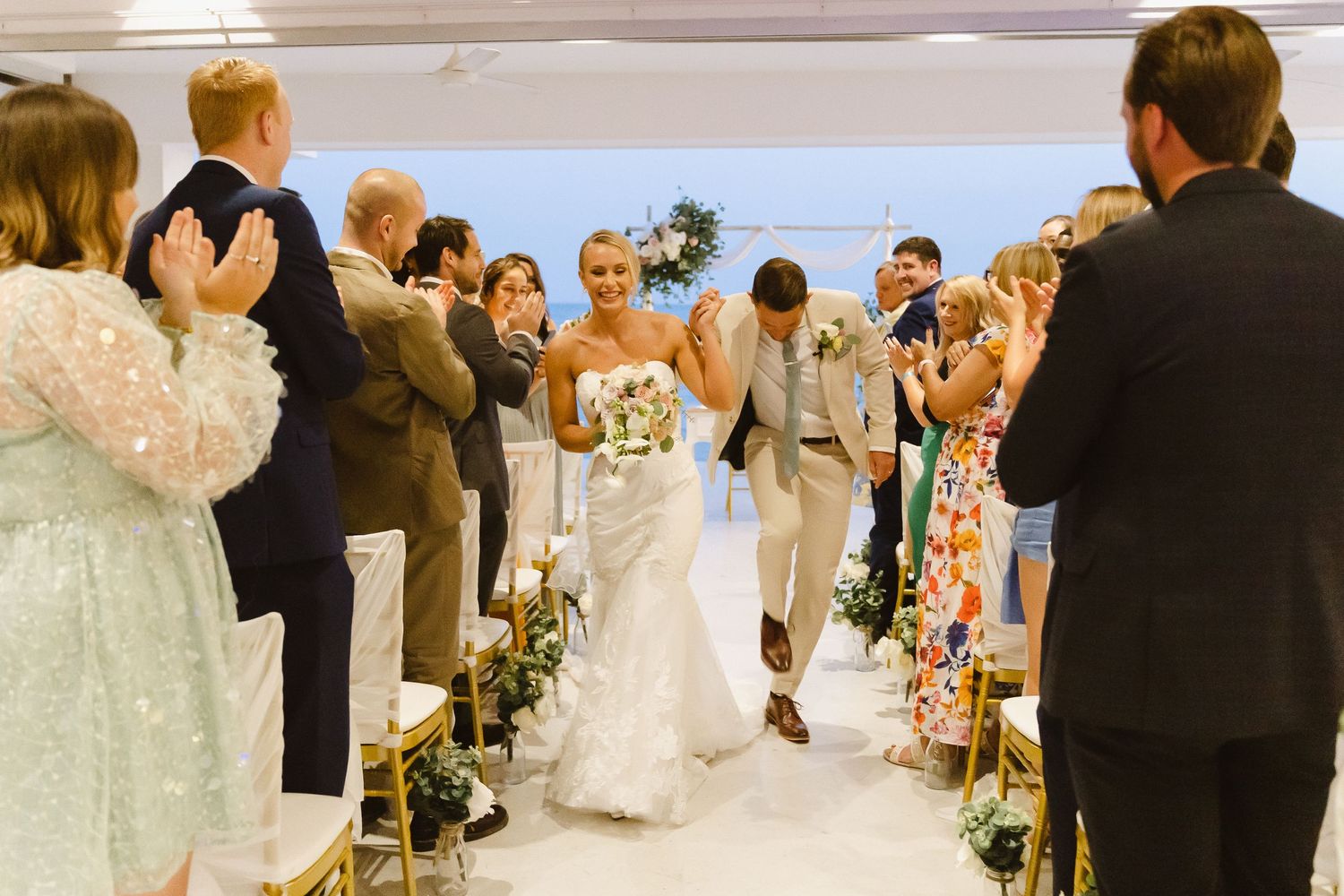 Happy couple walks down aisle while wedding guests cheer in bright indoor venue.