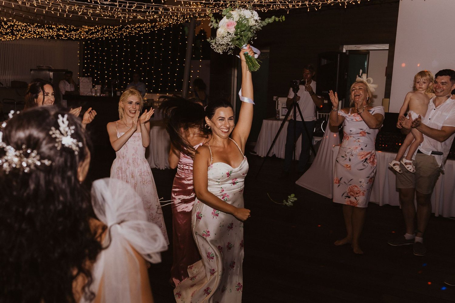 Wedding celebration reaches peak excitement during bouquet toss under glowing light canopy.