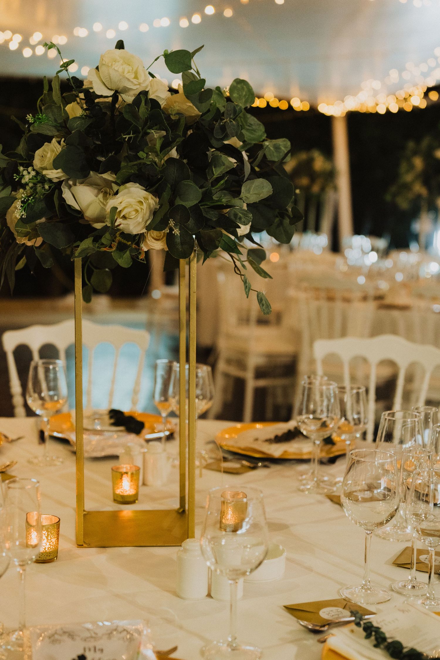 Modern wedding table centerpiece featuring white roses and greenery in a tall gold geometric vase.