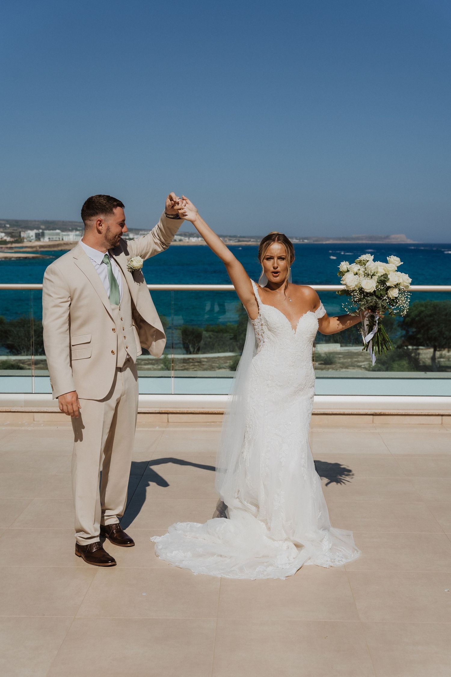 Newlywed couple dances together on a scenic balcony overlooking bright blue ocean waters on their wedding day.