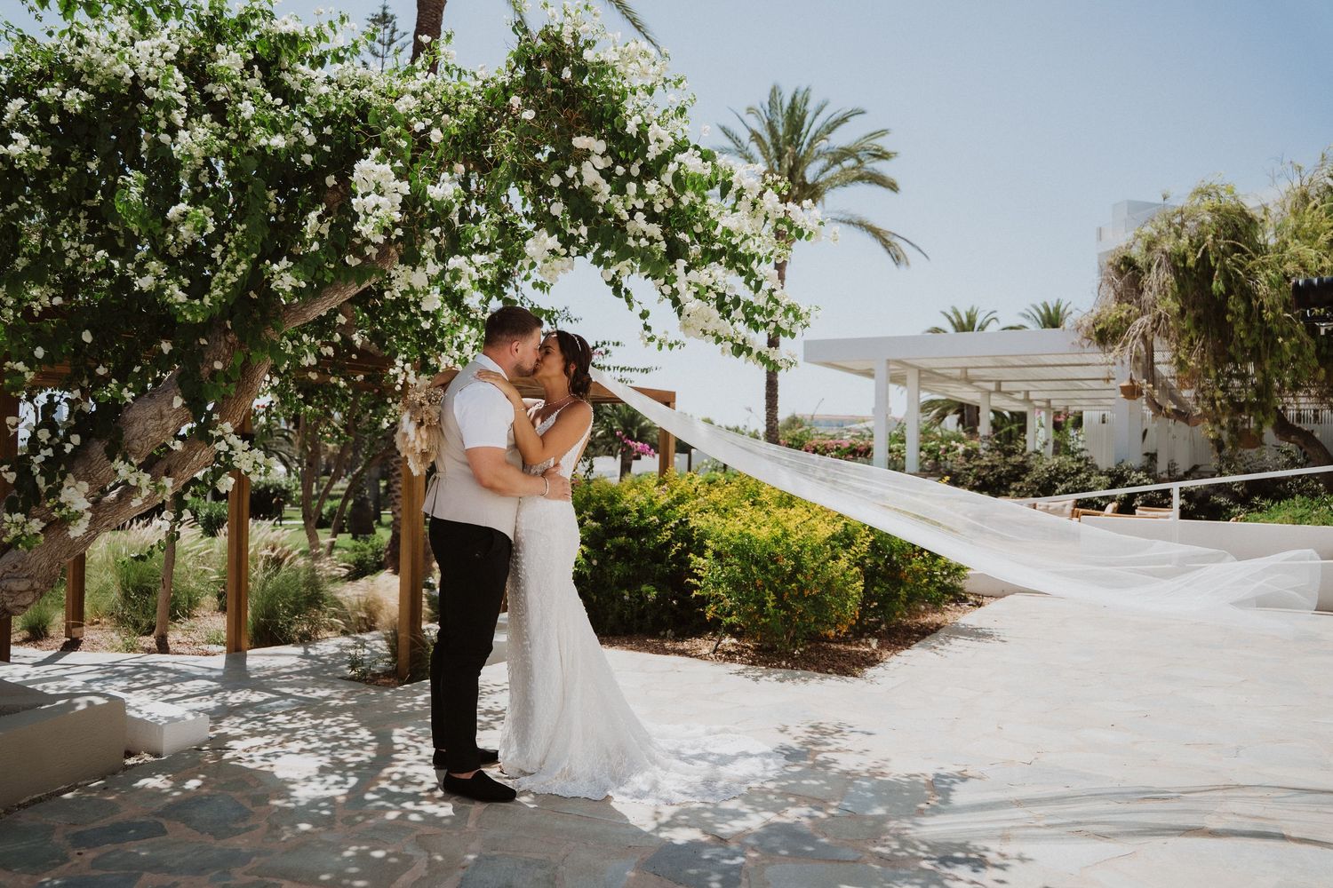 A couple shares an intimate moment under flowering trees at an elegant outdoor venue with palm trees and a white gazebo.