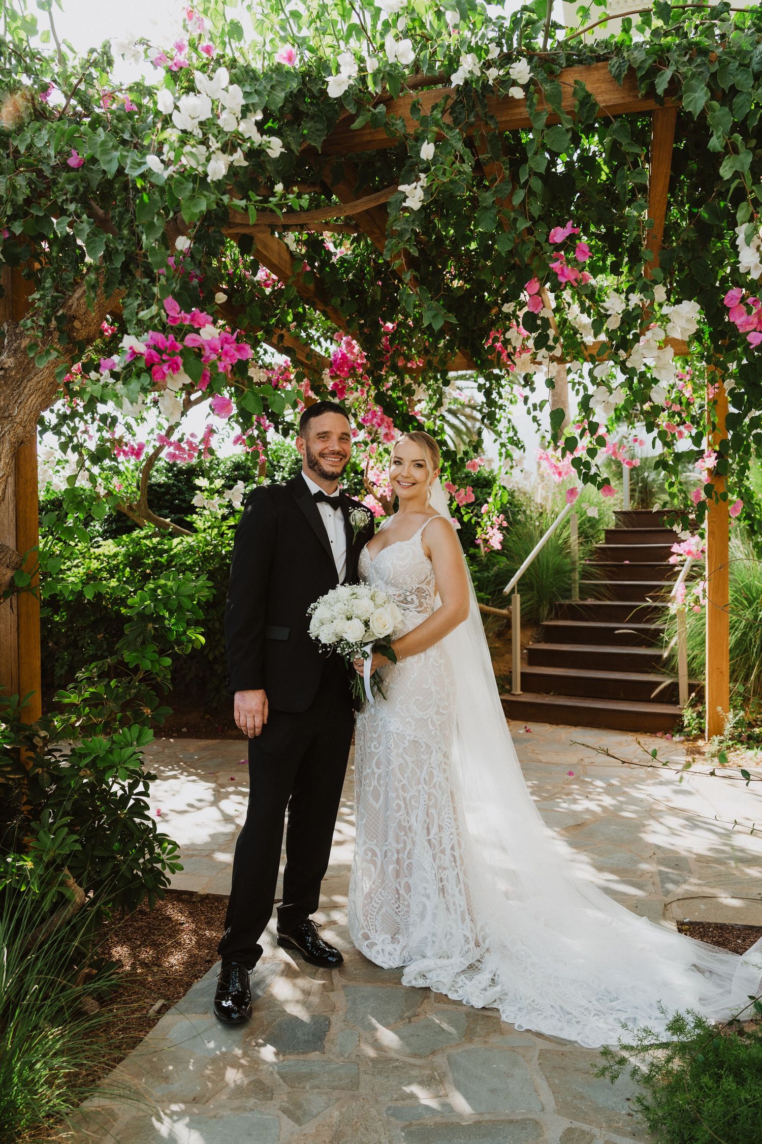 A couple poses under a wooden pergola covered in pink bougainvillea and white floral arrangements.