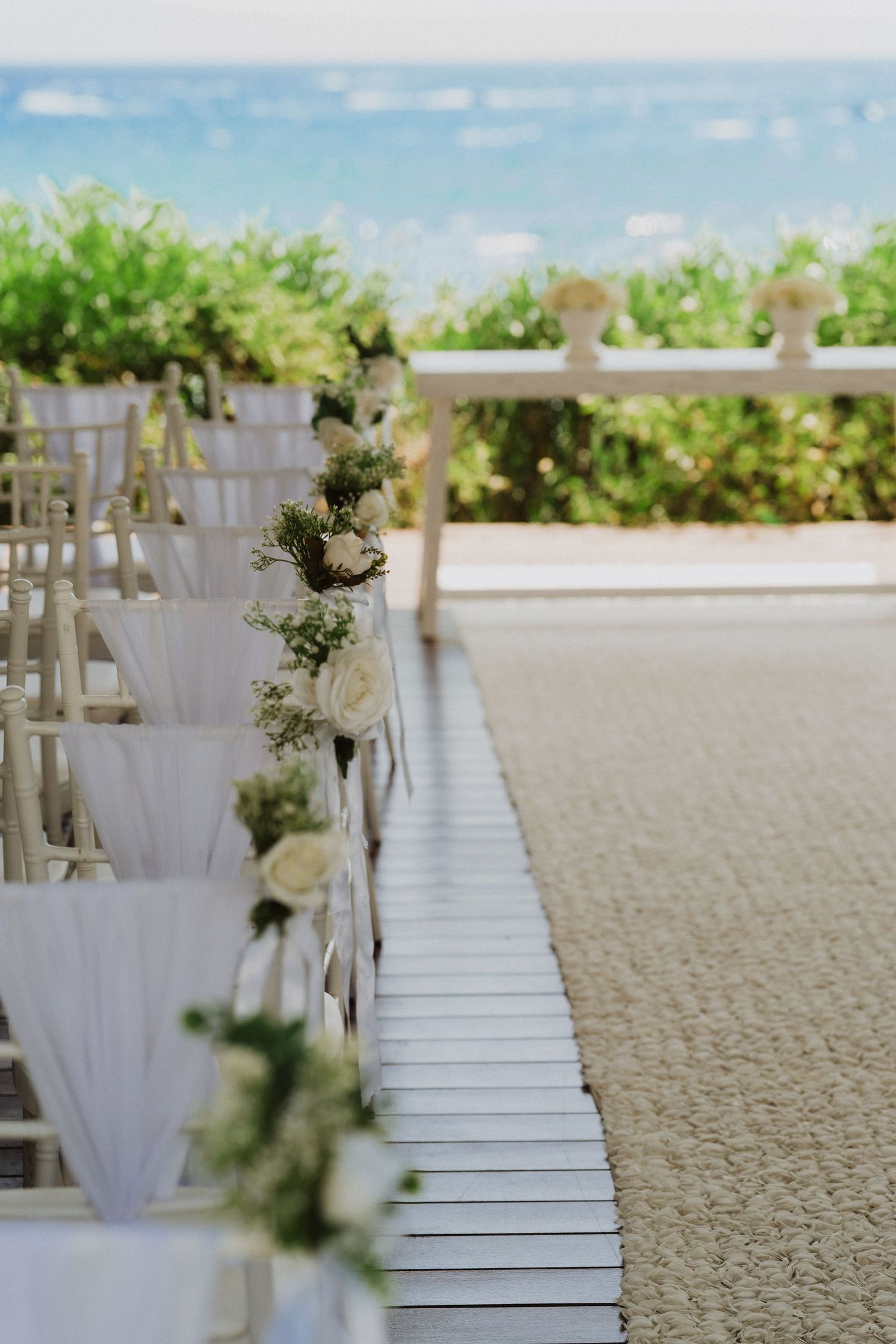 White wooden chairs line a beach aisle with small floral arrangements for an elegant seaside wedding ceremony setup.