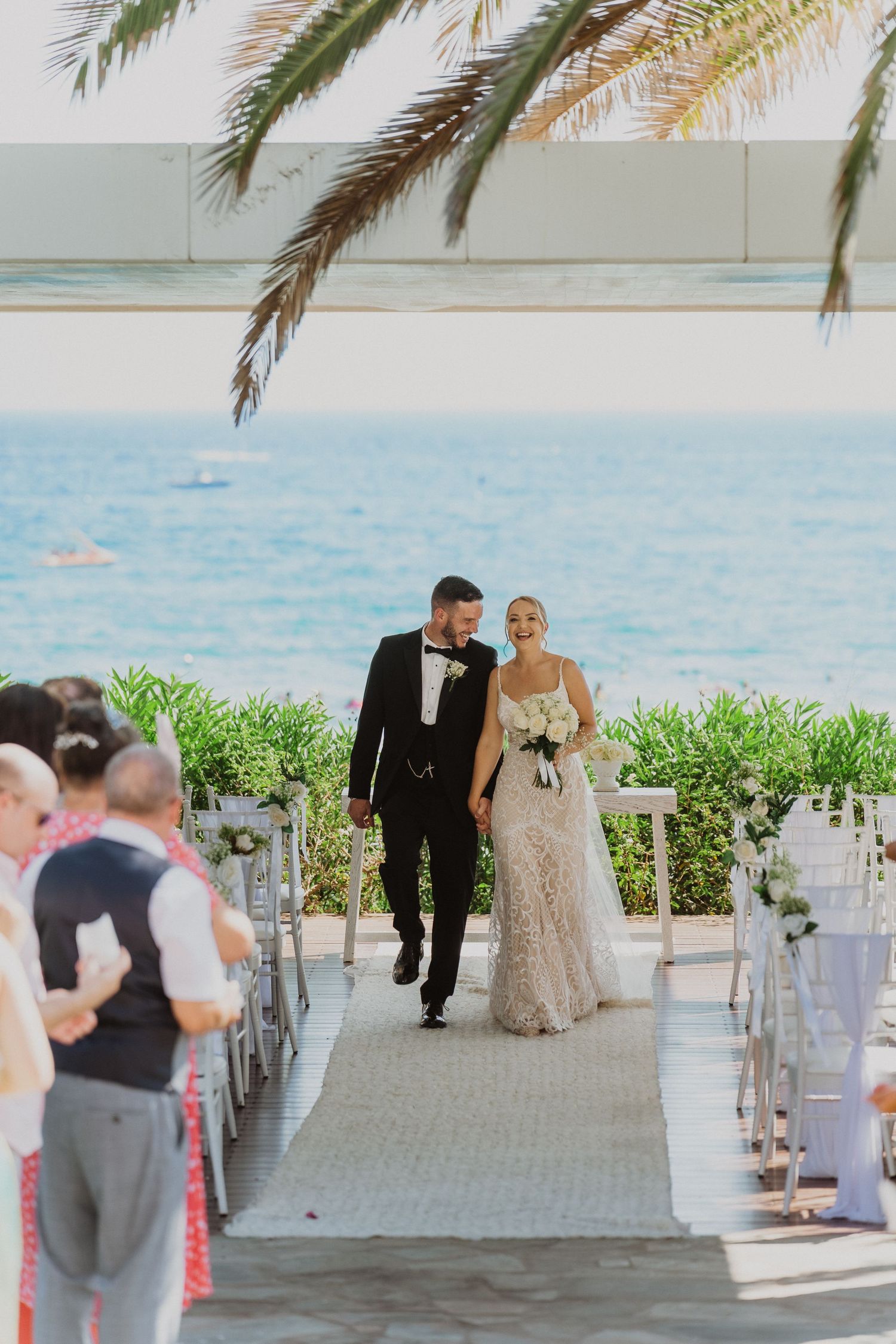 A couple walks down an aisle with palm trees and ocean views at a tropical beach wedding venue.