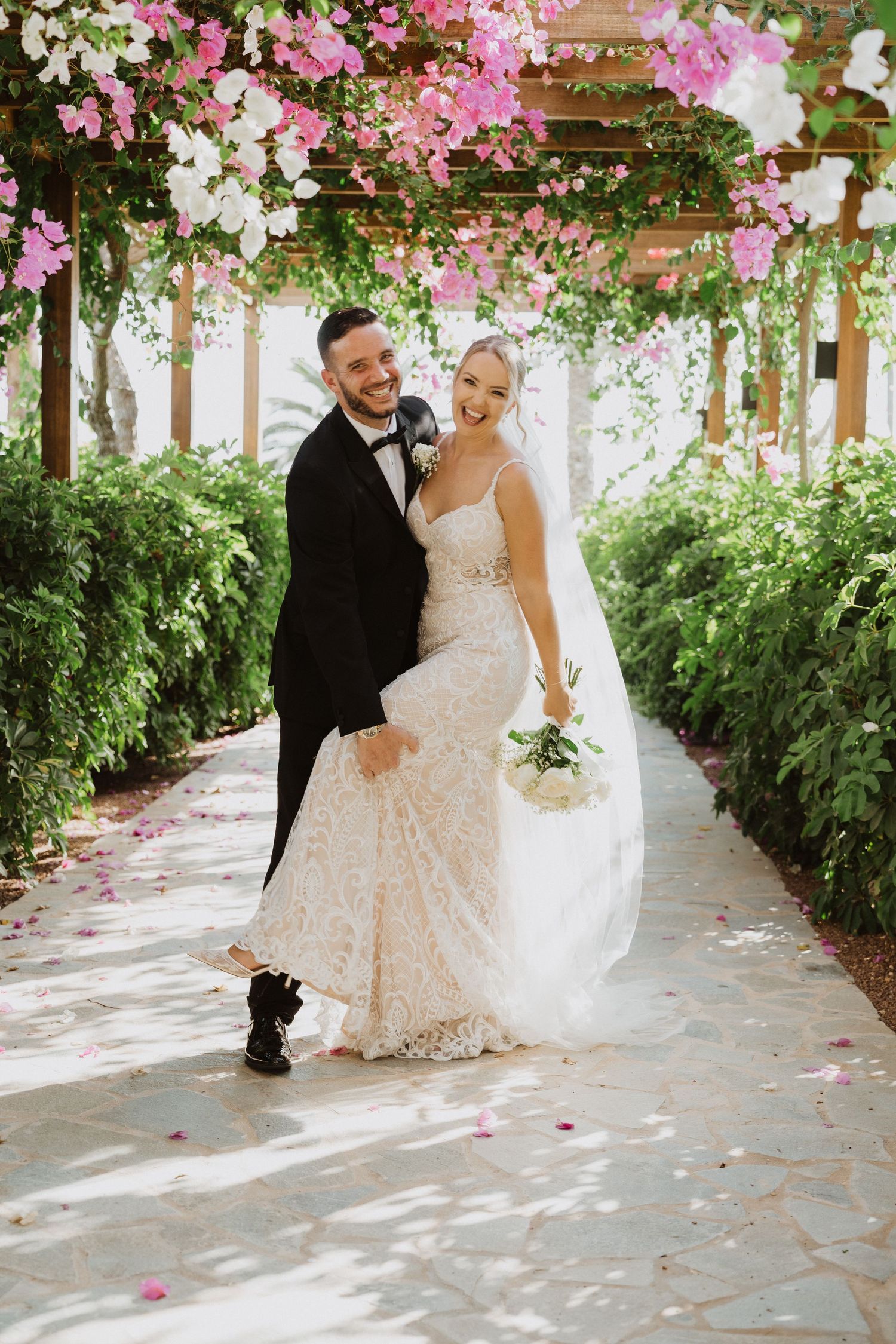 A romantic wedding photo under a beautiful pergola covered in pink and white flowering vines.