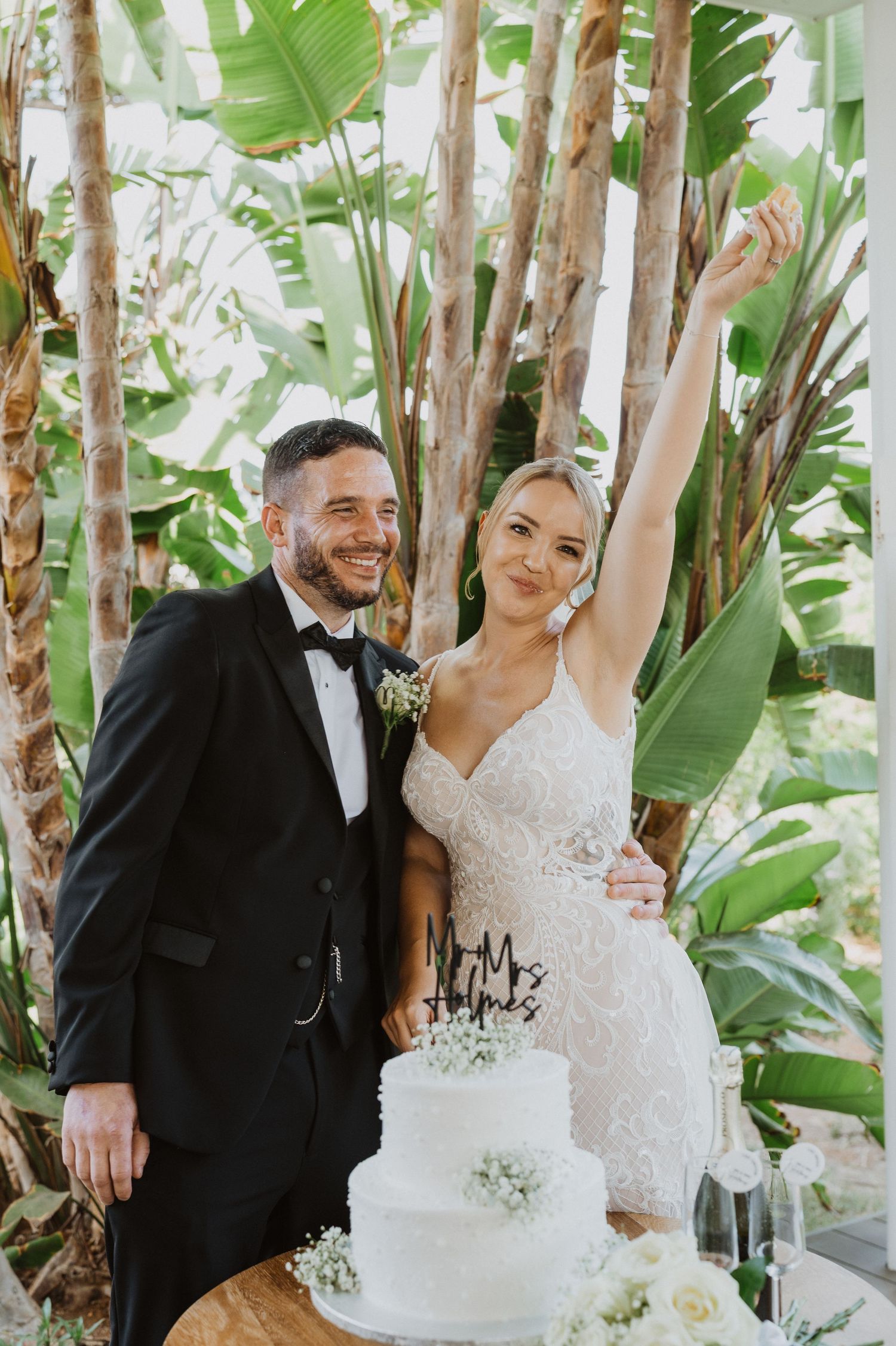 A joyful wedding couple shares a celebratory moment surrounded by tropical foliage and trees.