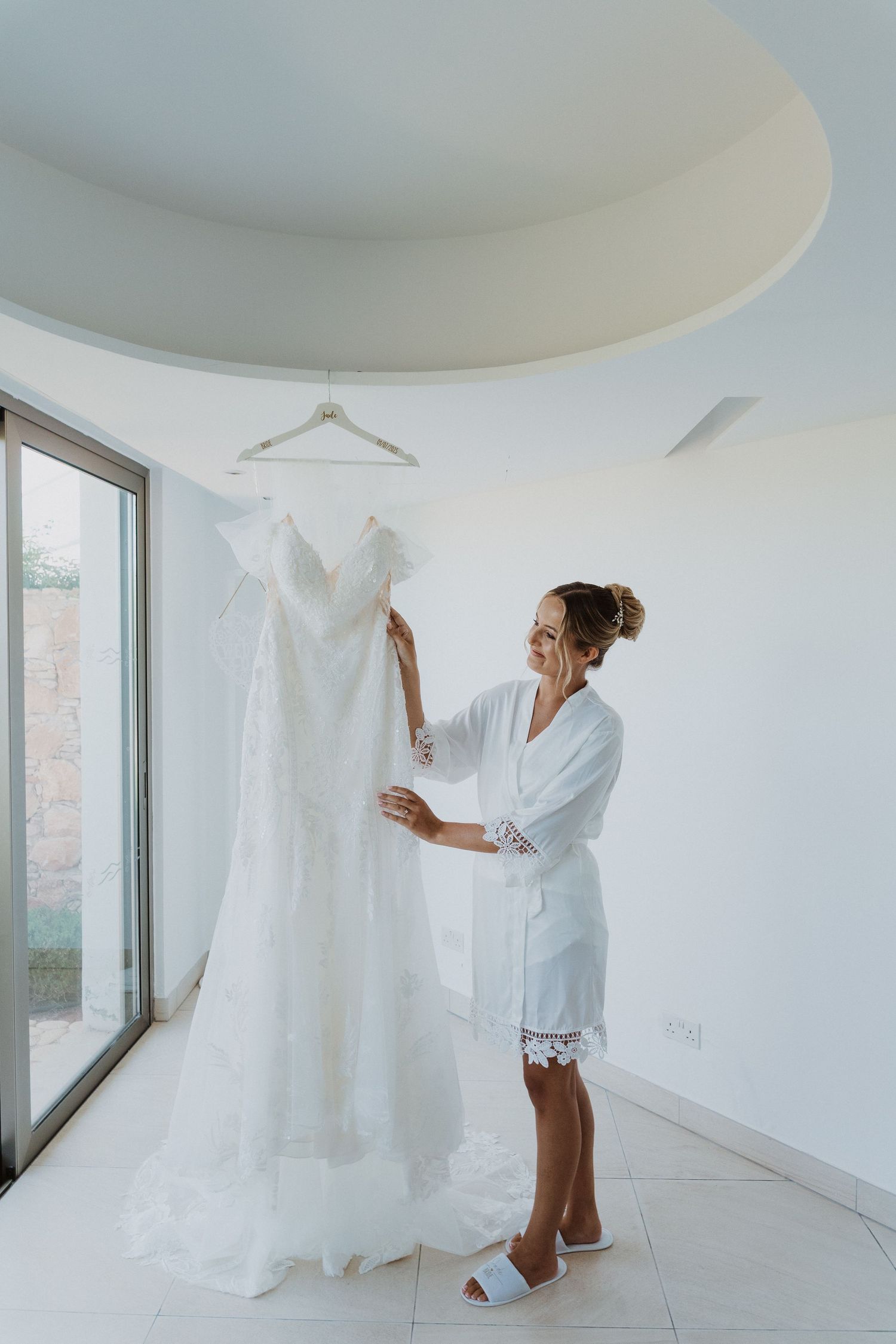 A white wedding dress hangs in a bright minimalist room while someone in a white robe stands nearby.