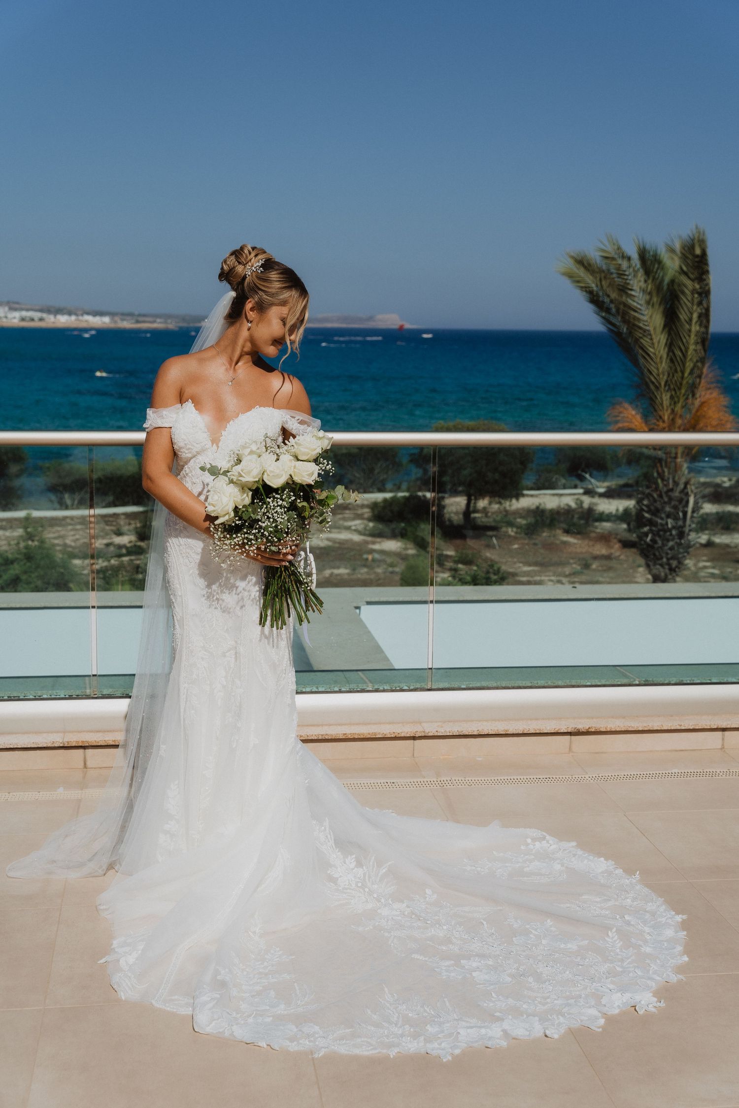 Bride in flowing lace wedding dress with long train stands on oceanfront terrace with palm trees and blue waters.
