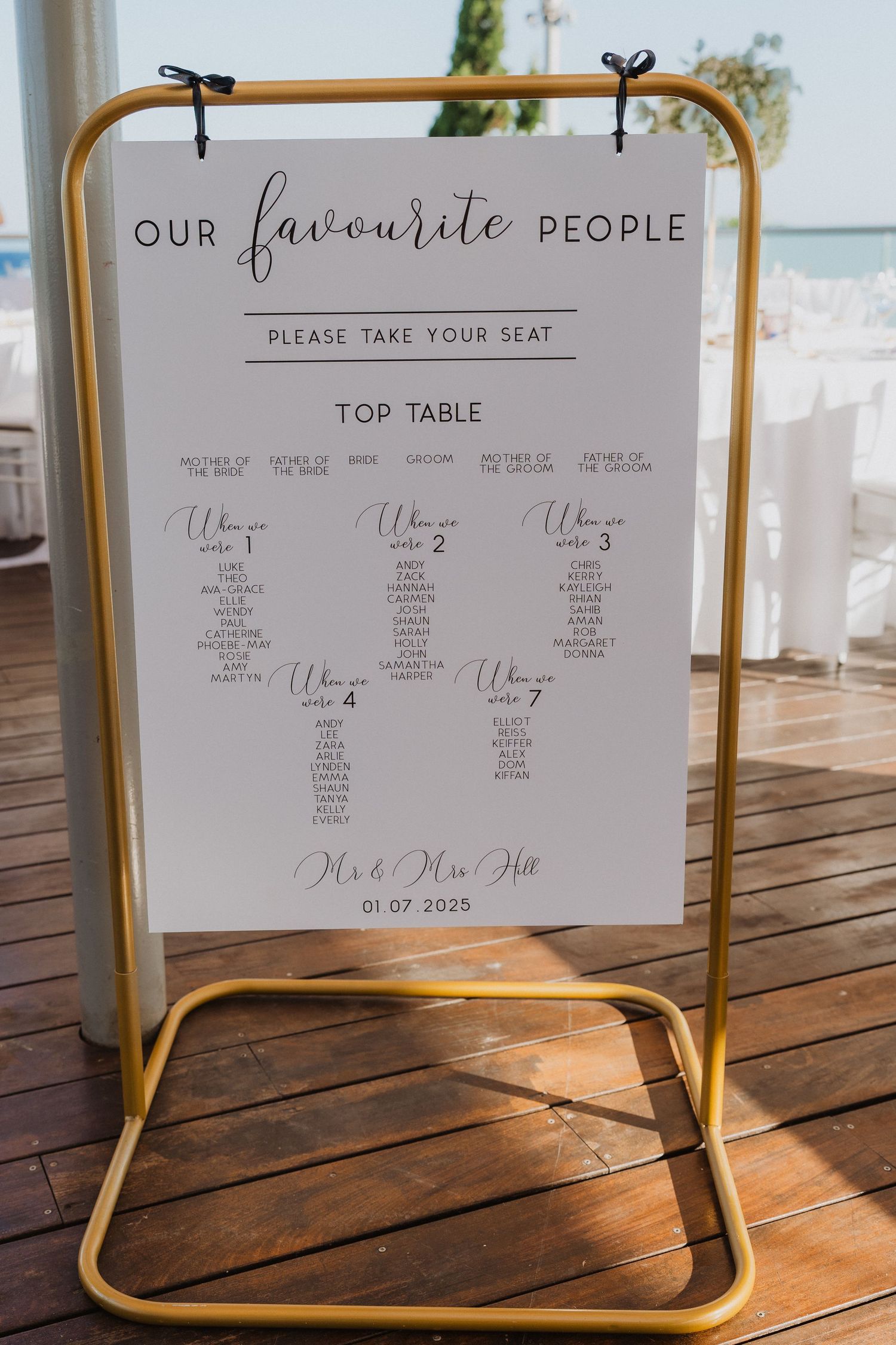 Wedding seating chart signs displayed on gold metal stands at an outdoor venue with ocean views in the background.