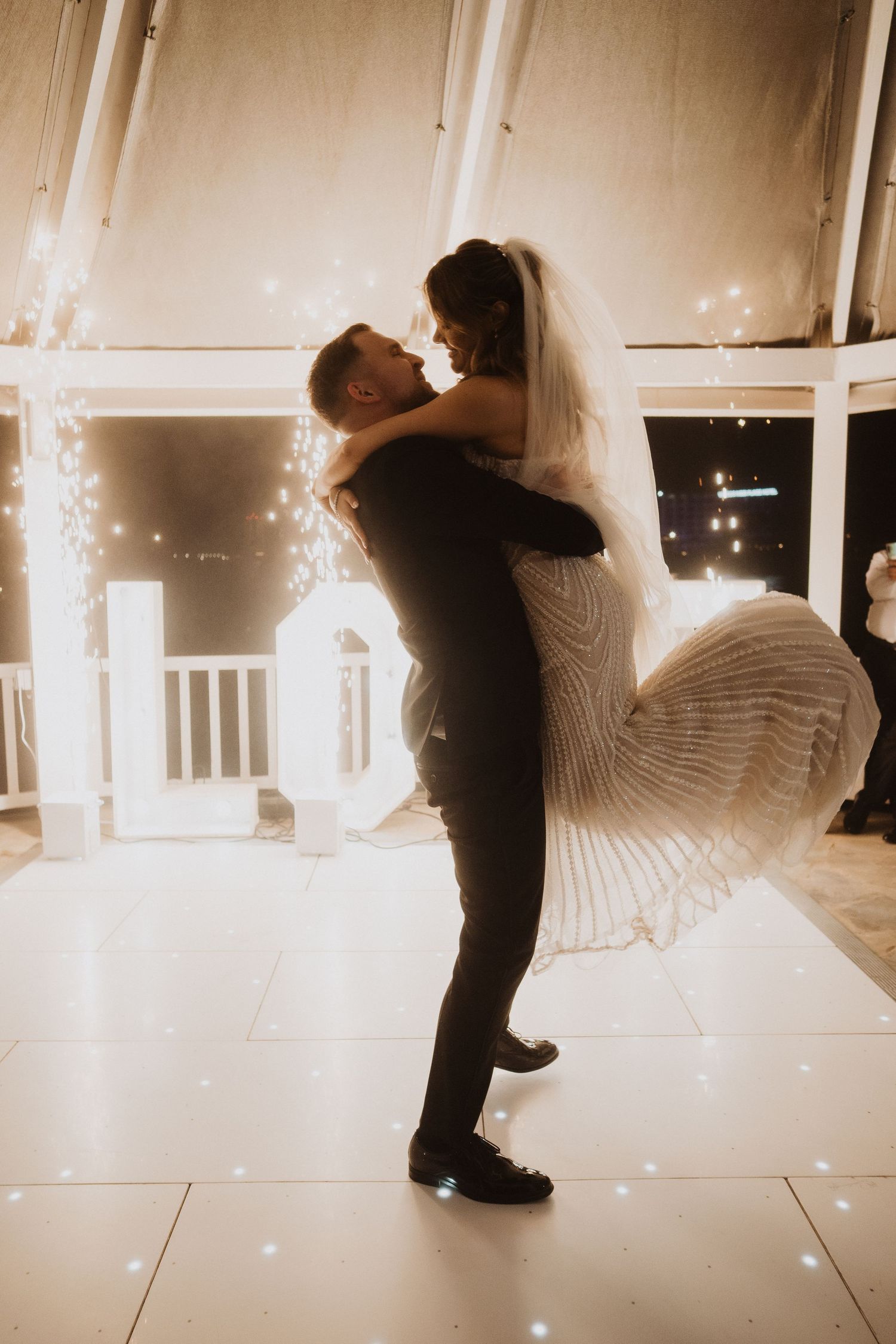 Groom lifting bride in wedding dress during romantic first dance with sparkling LOVE sign in background.