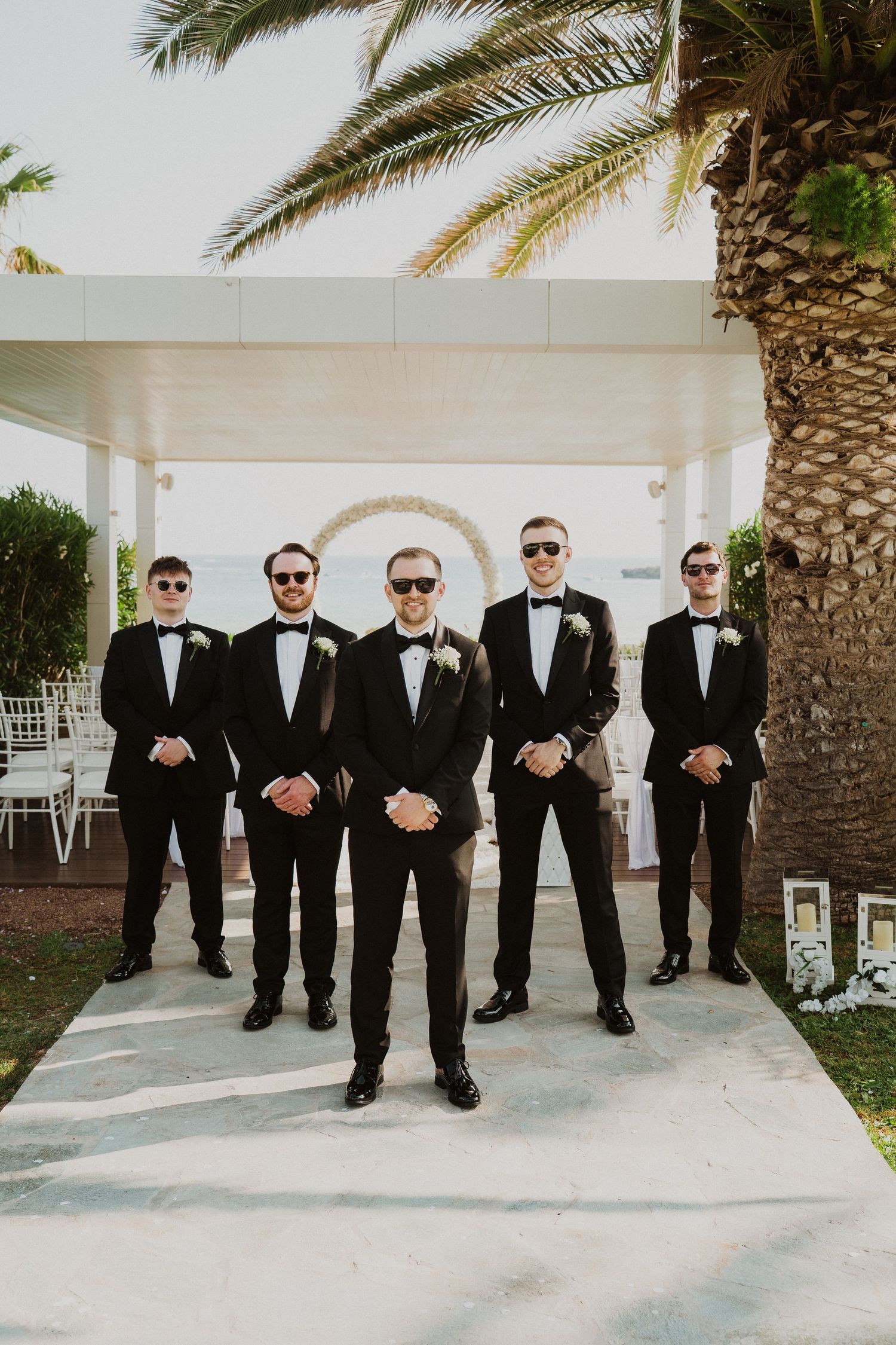 Five groomsmen in matching black tuxedos and sunglasses pose at outdoor wedding venue.