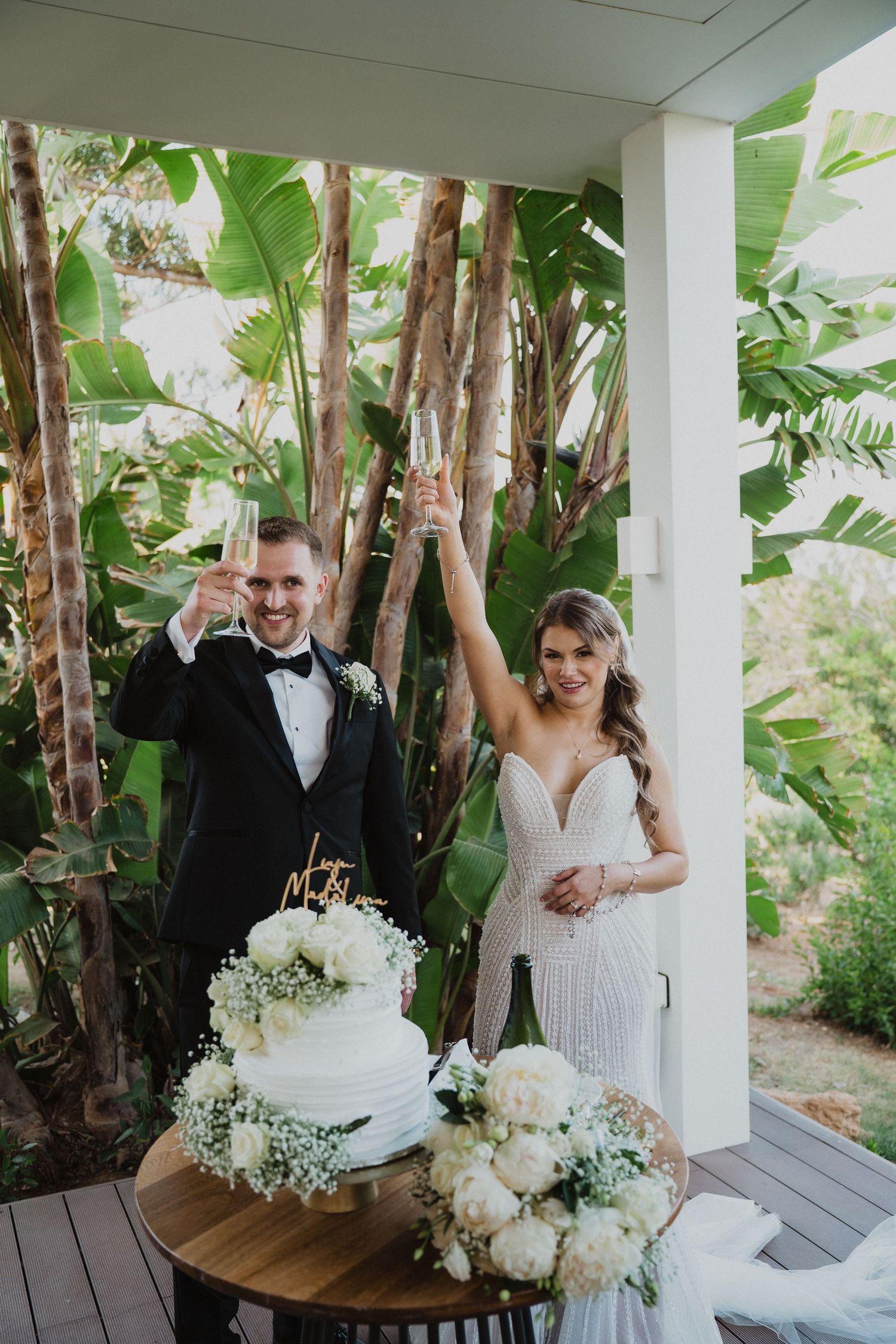 Bride and groom toast champagne beside a white wedding cake decorated with white roses and baby's breath.