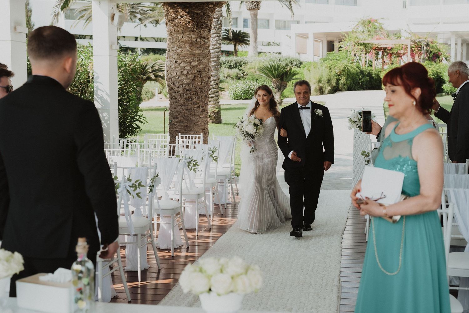 Bride and father walking down outdoor wedding ceremony aisle toward waiting groom and guests.