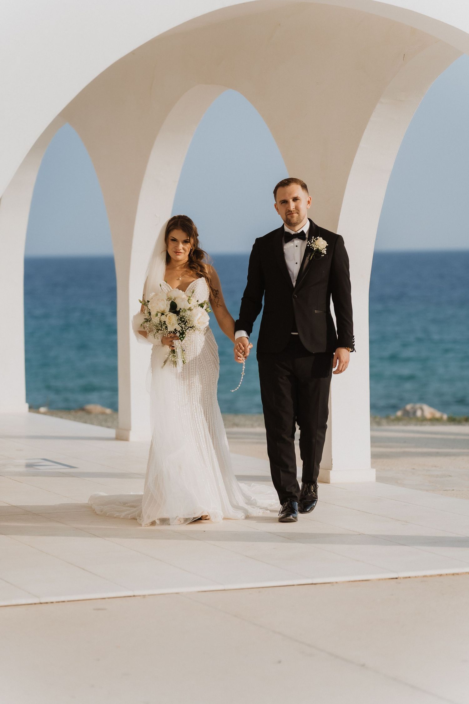 Bride and groom walk hand-in-hand through white arched colonnade with blue ocean backdrop at seaside wedding.
