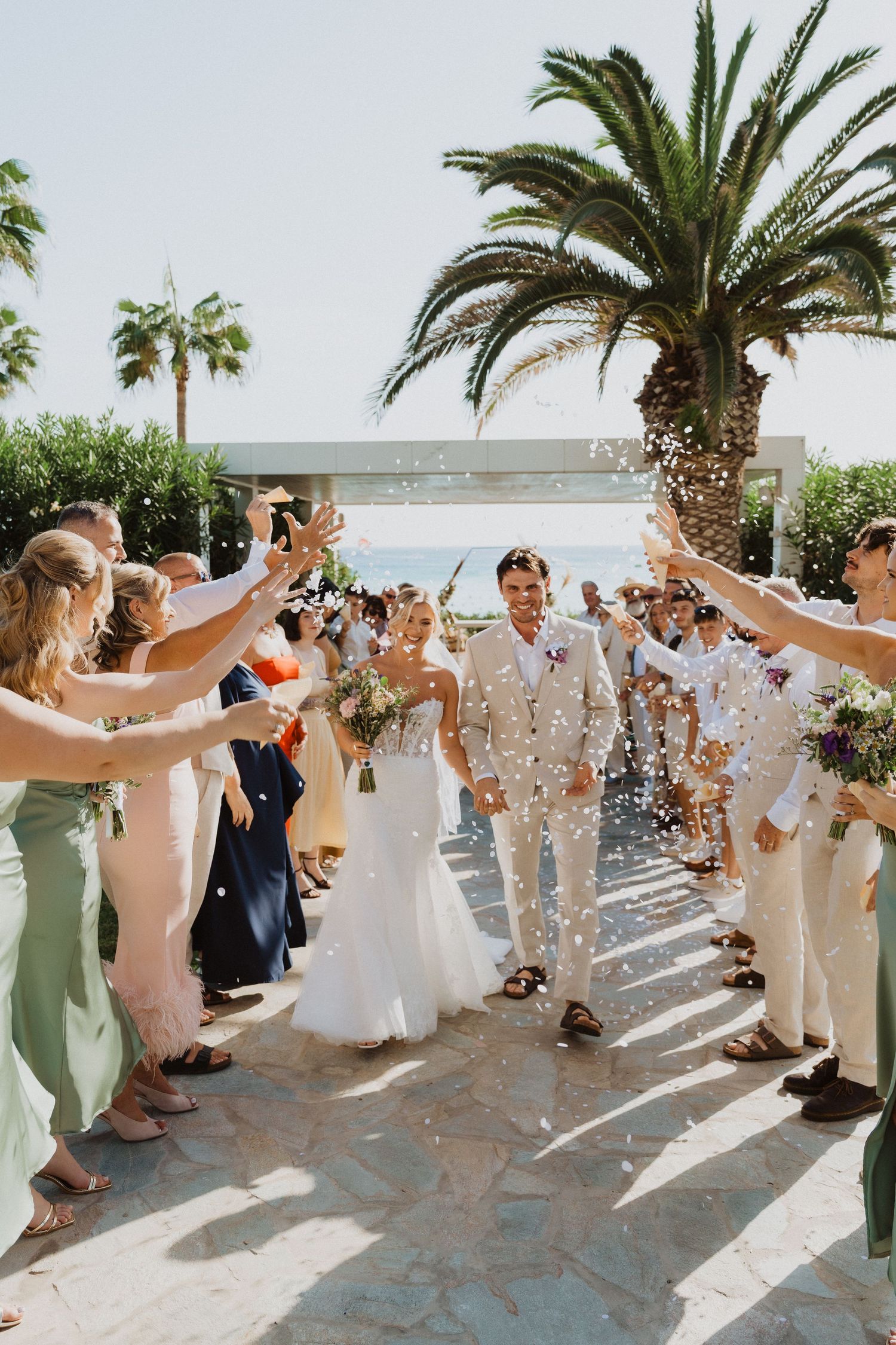 Guests line up to throw confetti during a joyous wedding celebration with palm trees in the background.