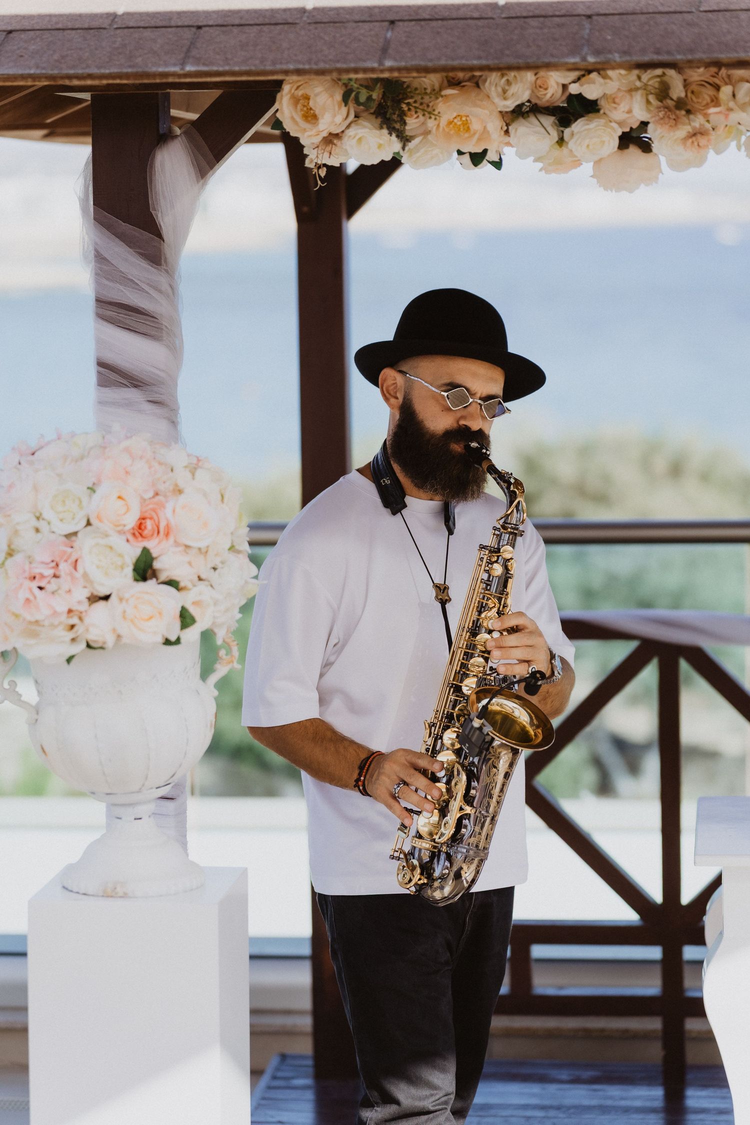 Saxophone player in a hat performing next to floral arrangements at an outdoor venue.