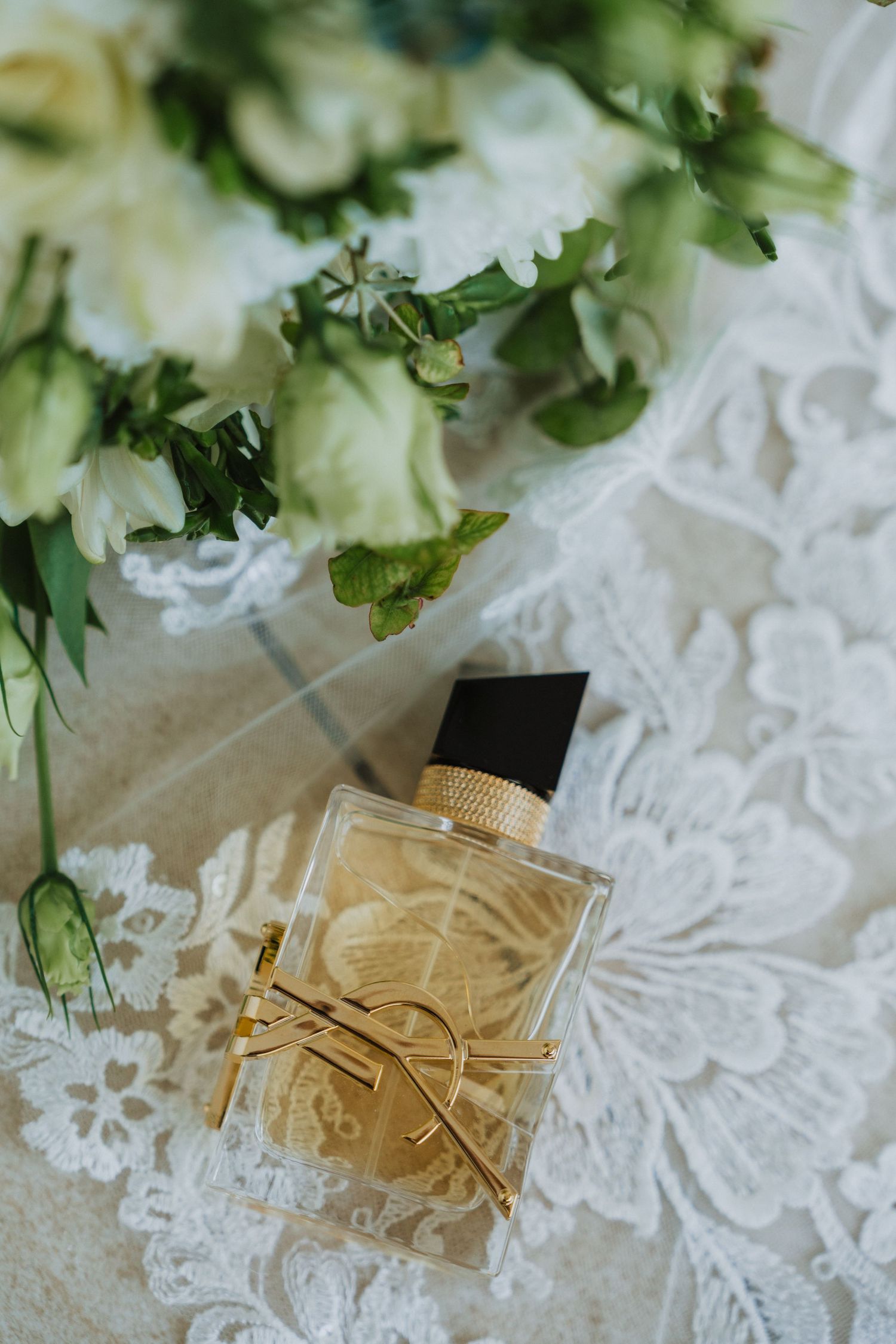 Wedding rings and perfume bottle arranged on delicate white lace with greenery accents.